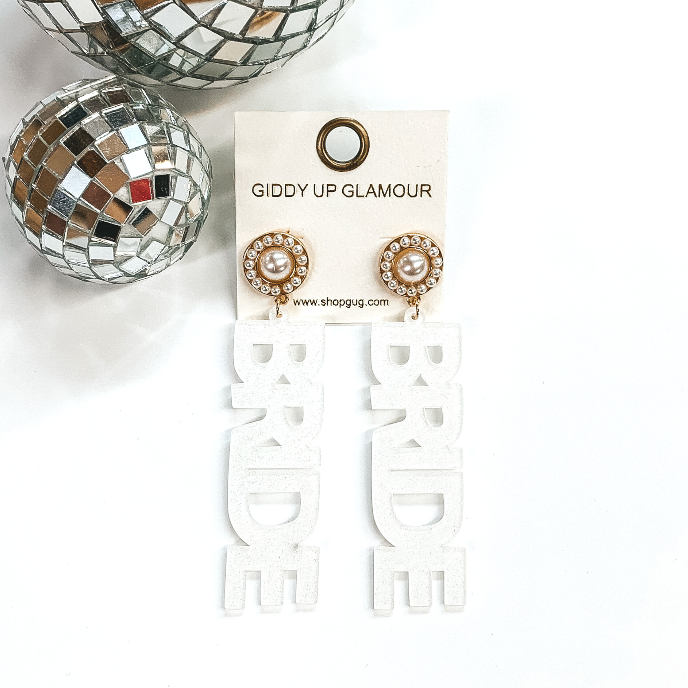Gold circle studs with white pearls. There is a drop pendant in white glitter that spells out "BRIDE." These earrings are pictured on a white background with disco balls in the top left corner. 