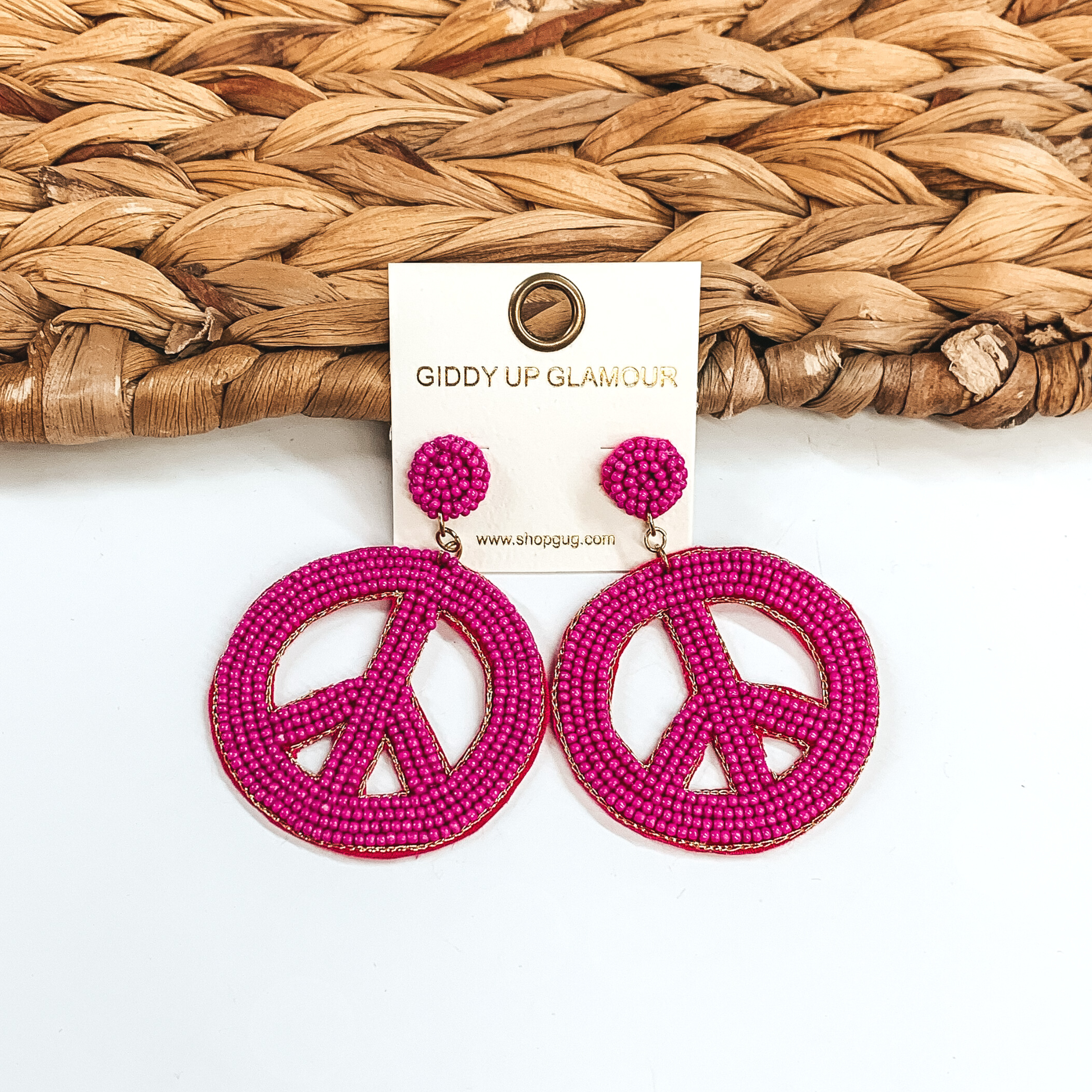 Circle beaded stud earrings with a beaded peace sign in fuchsia. These earrings are pictured on a half white and half basket weave background.