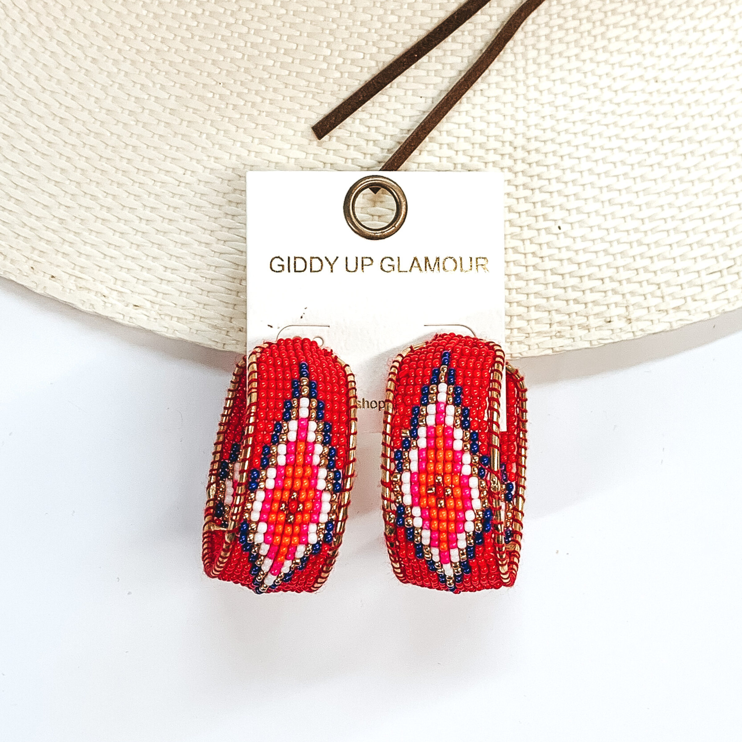Beaded hoop earrings in red with an aztec design in multicolor. These earrings are pictured on a white background on a straw hat. 
