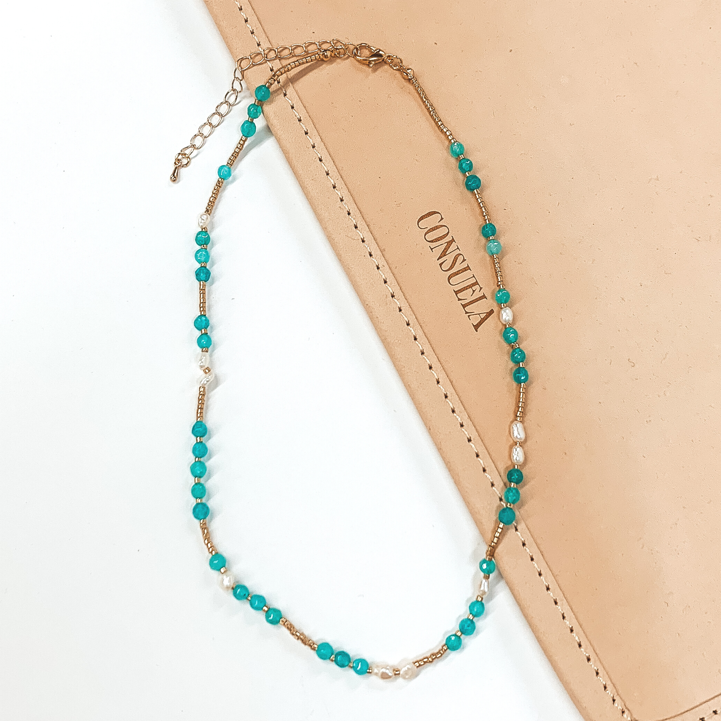 This necklace include gold beads, white pearl beads, and turquoise crystal beads. This necklace is pictured on a white and tan background. 