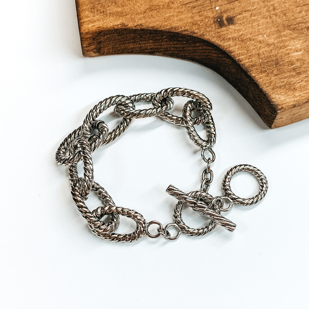 Twisted gold chain bracelet with toggle clasp in silver. This bracelet is pictured on a white background with brown wood in the corner. 