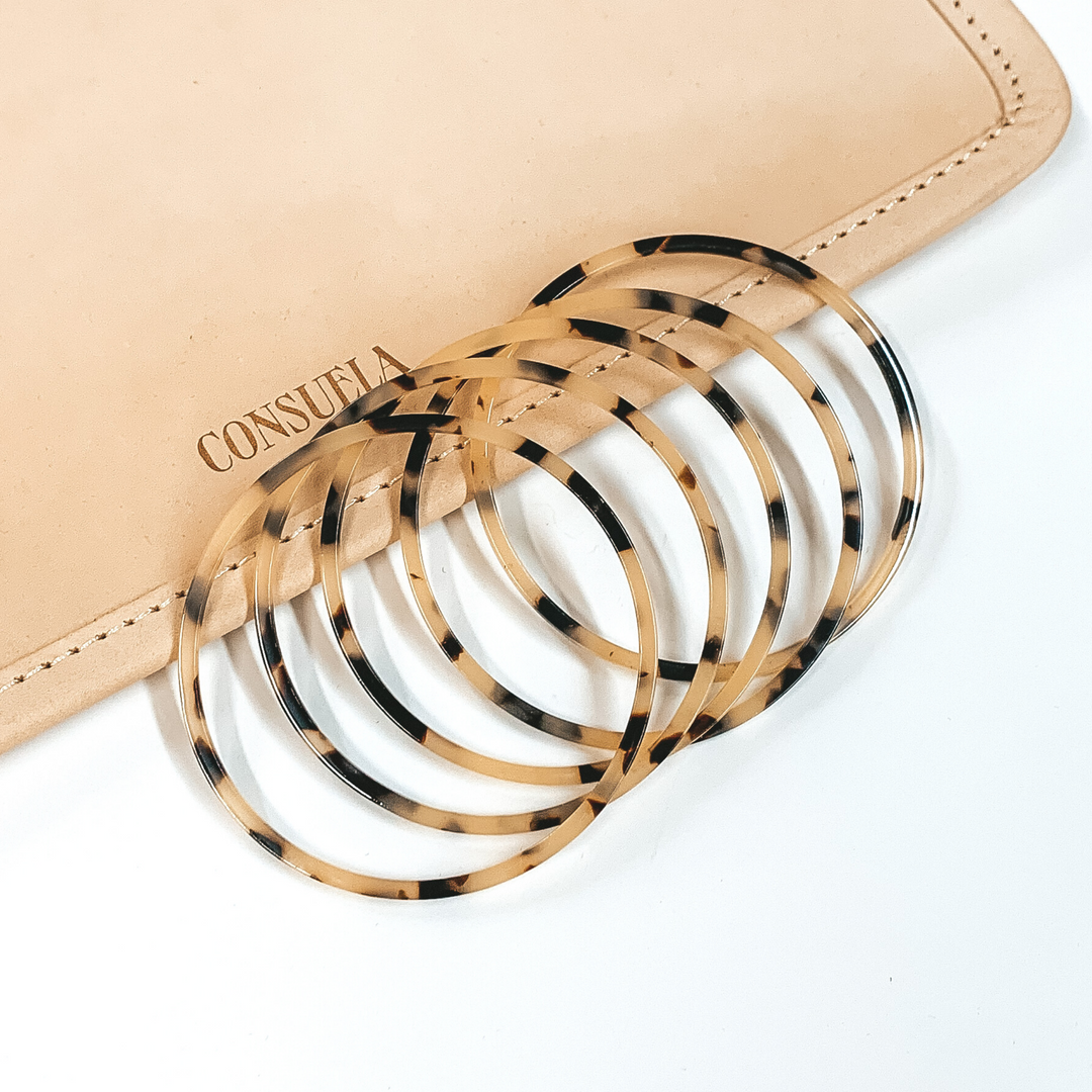 Five tortoise print bracelets partially laying on a white and tan background.