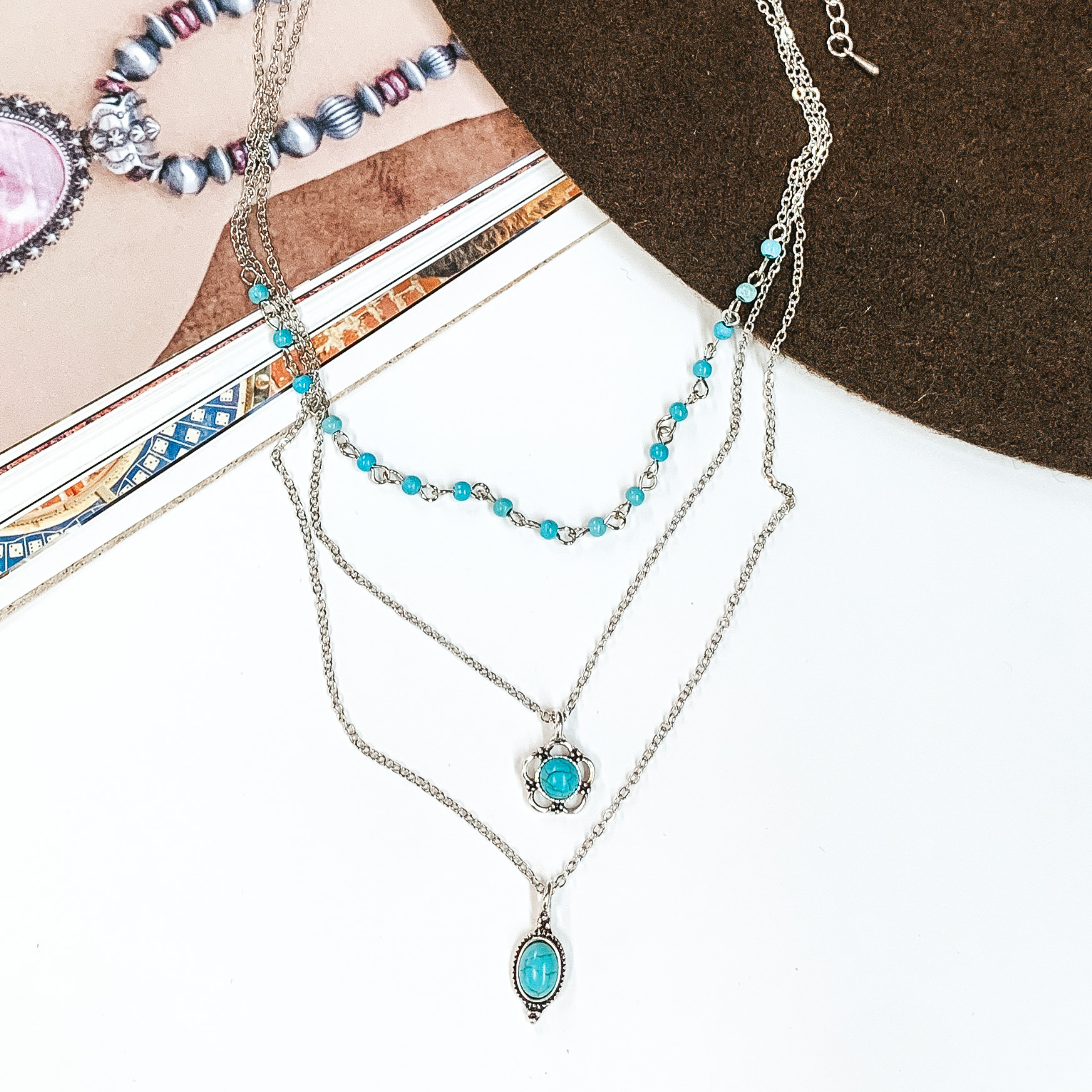 Triple layered, silver, chain necklaces. Two strands have a small turquoise stone pendant with a silver outlining and the shortest chain has a turquoise beaded section. This necklace is pictured partially on an open magazine on a white background. 