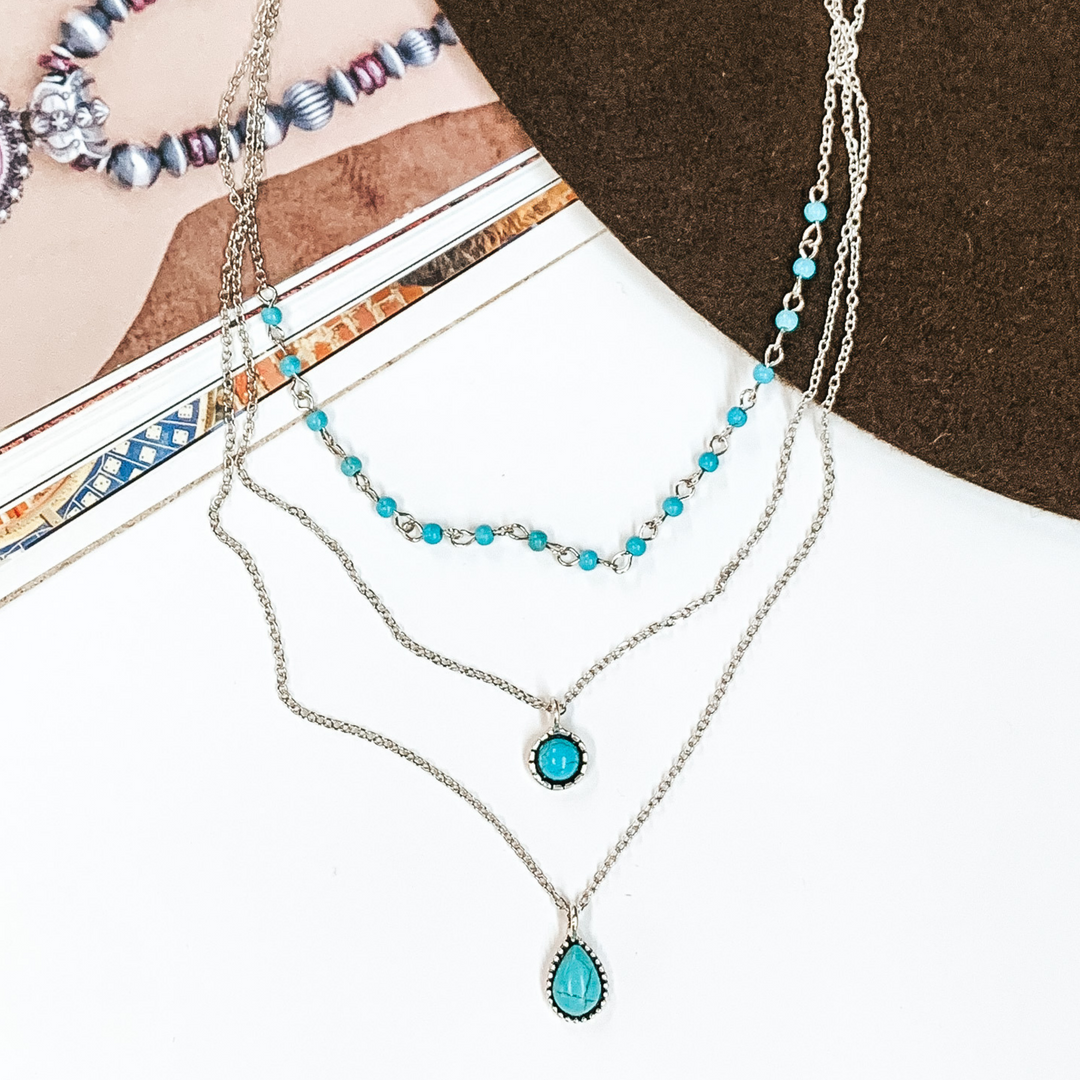 Triple layered, silver, chain necklaces. Two strands have a small turquoise stone pendant with a silver outlining and the shortest chain has a turquoise beaded section. This necklace is pictured partially on an open magazine on a white background. 