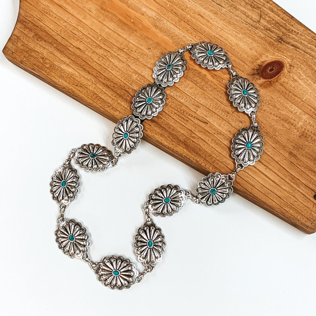 Silver, oval concho hat band with small turquoise stones in the middle of each concho. This hat band is pictured partially laying on a brown block on a white background. 
