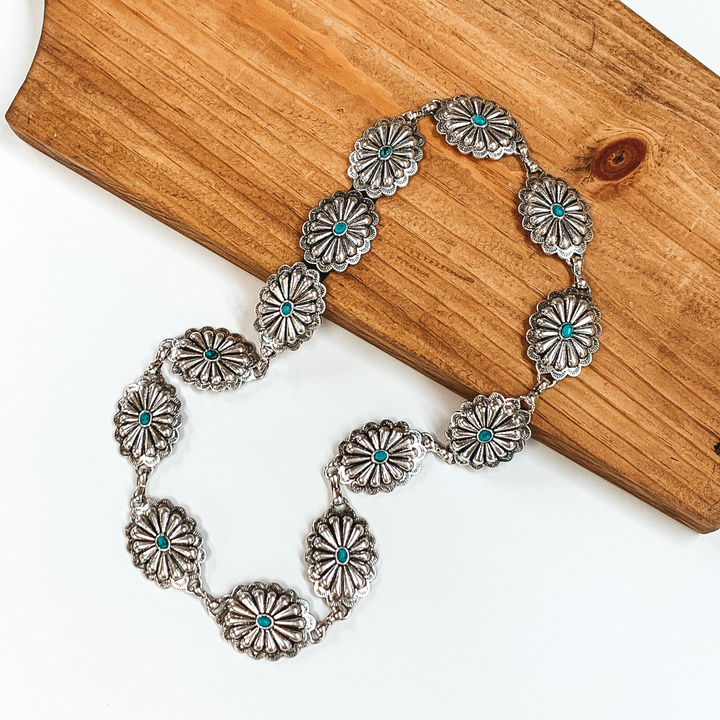 Silver, oval concho hat band with small turquoise stones in the middle of each concho. This hat band is pictured partially laying on a brown block on a white background. 