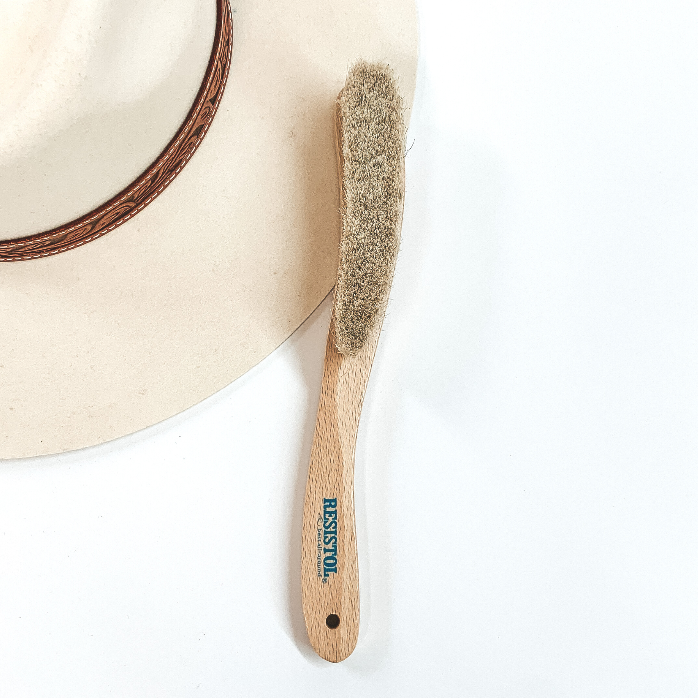 Thin wooden brush with ivory bristles. This brush is pictured laying partially on an ivory hat on a white background. 