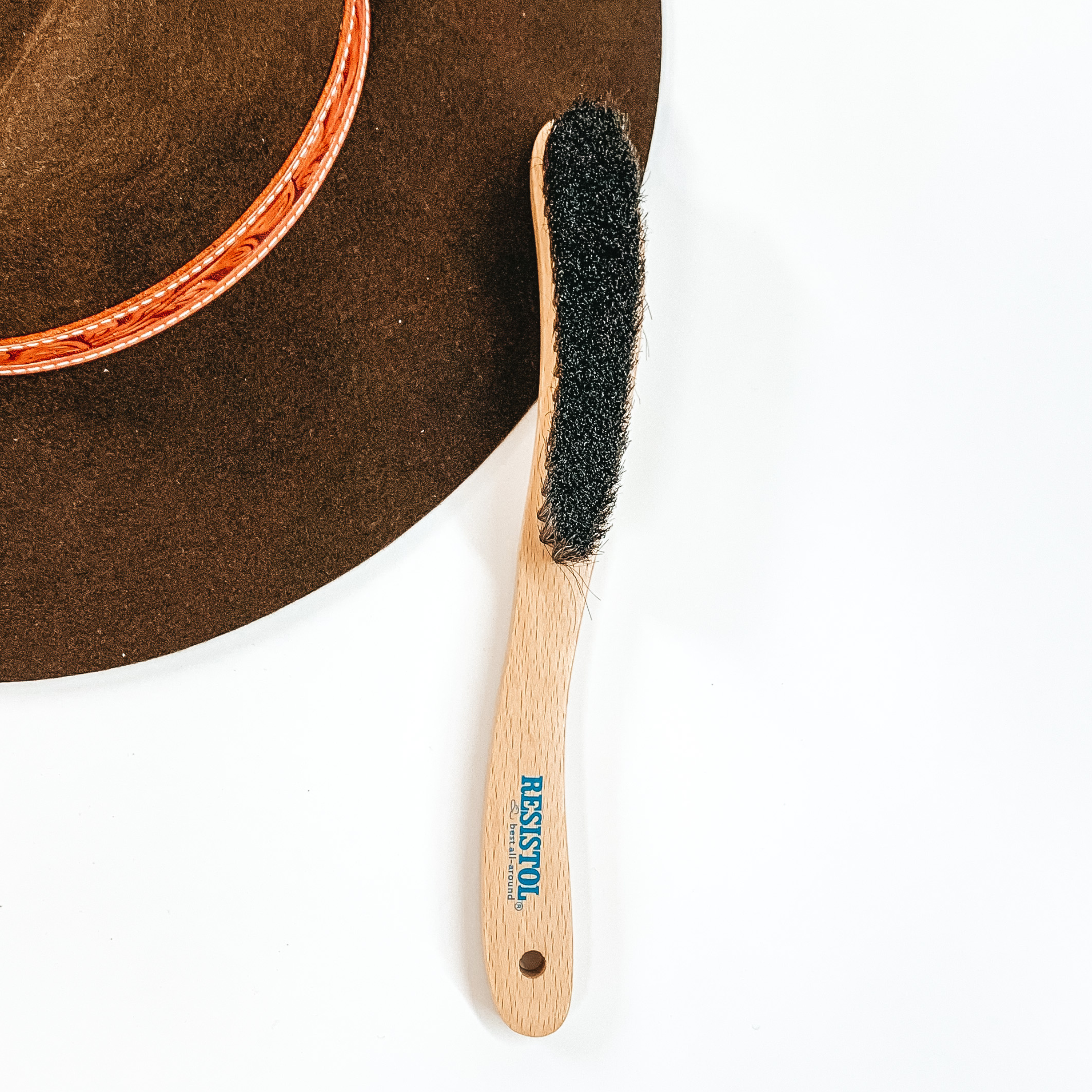 Thin wooden brush with brown bristles. This brush is pictured laying partially on a brown hat on a white background. 