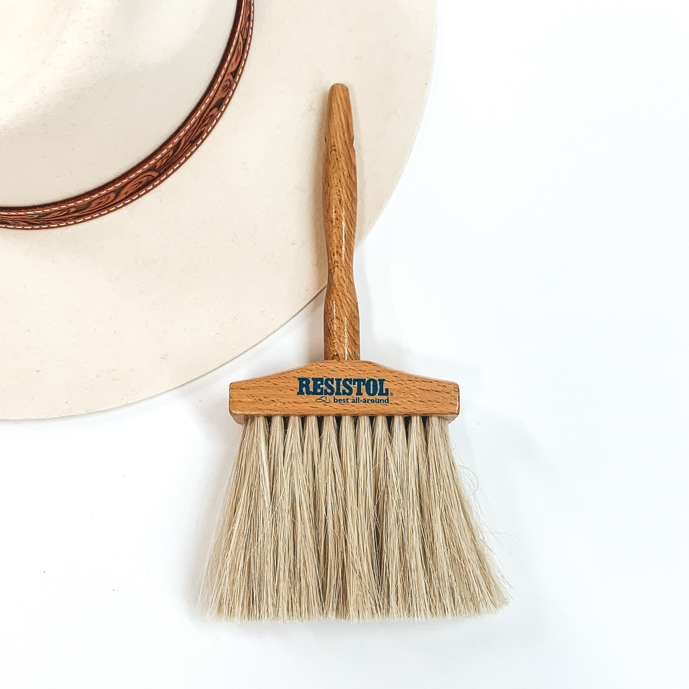 Wide, ivory bristle brush with a wooden handle, This brush is pictured partially laying on an ivory hat on a white background. 