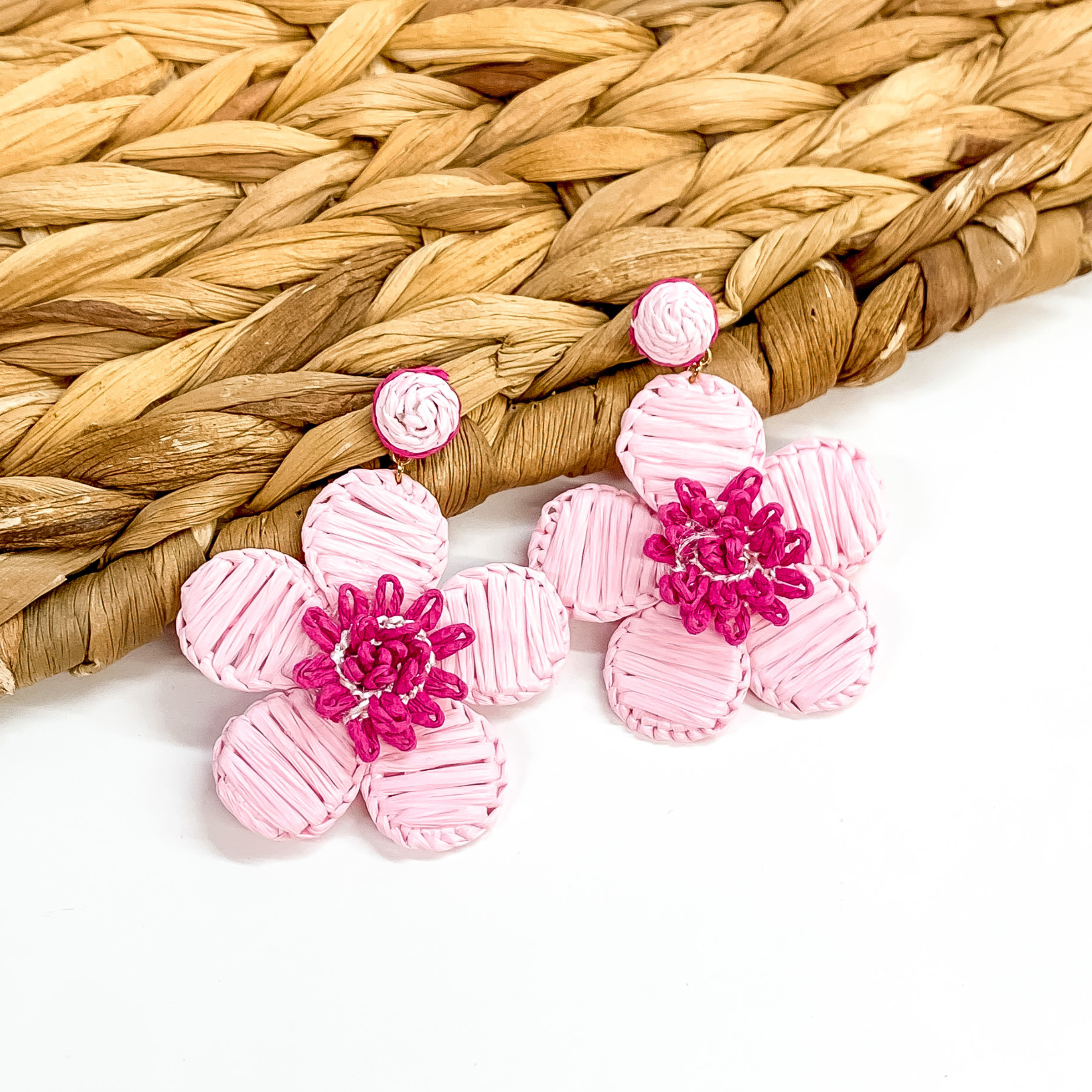 Baby pink affia wrapped circle studs with hanging flower pendant wrapped in baby pink  raffia and fuchsia colored middle. These earrings are pictured partially laying on a basket weave material on a white background. 