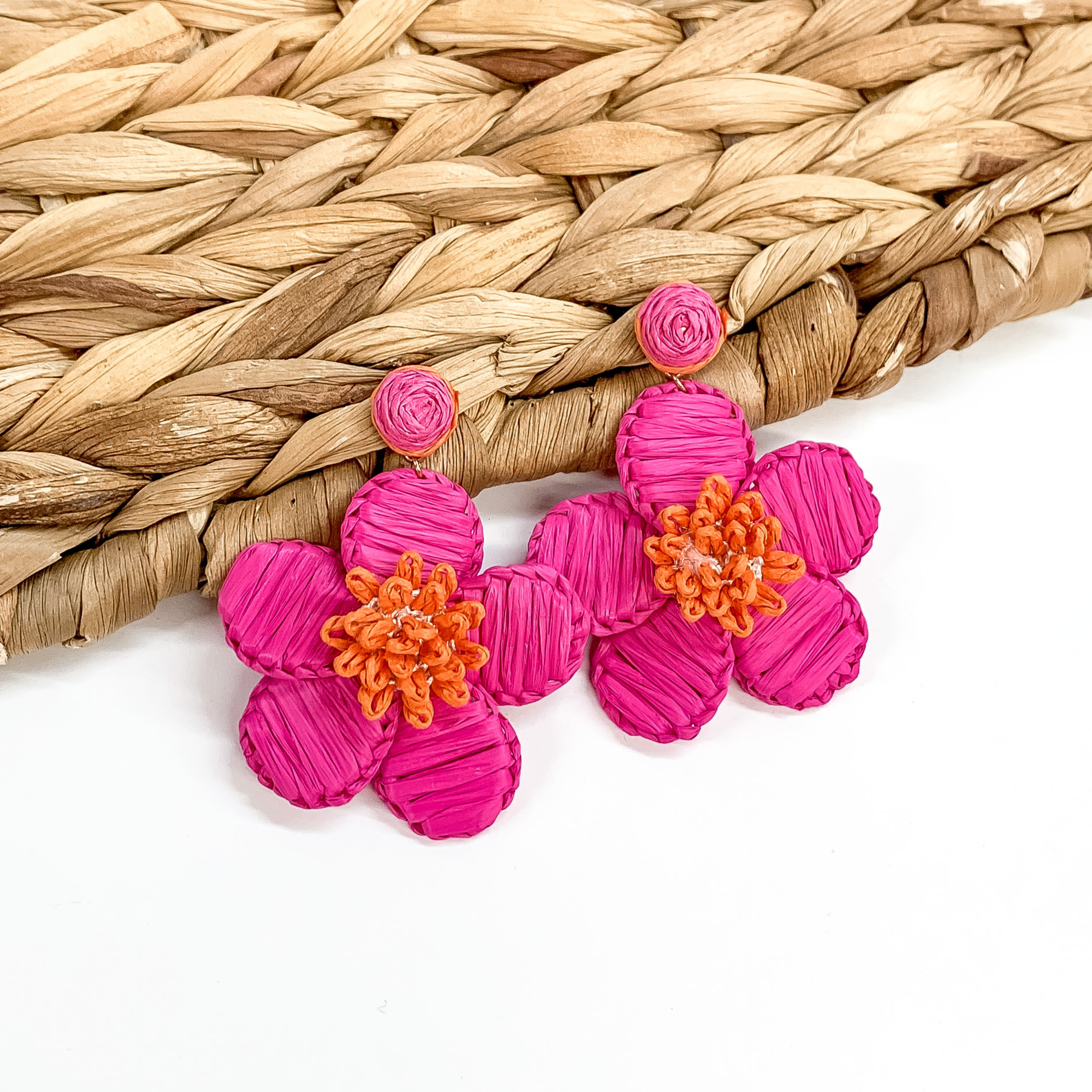 Hot pink affia wrapped circle studs with hanging flower pendant wrapped in hot pink  raffia and orange colored middle. These earrings are pictured partially laying on a basket weave material on a white background. 