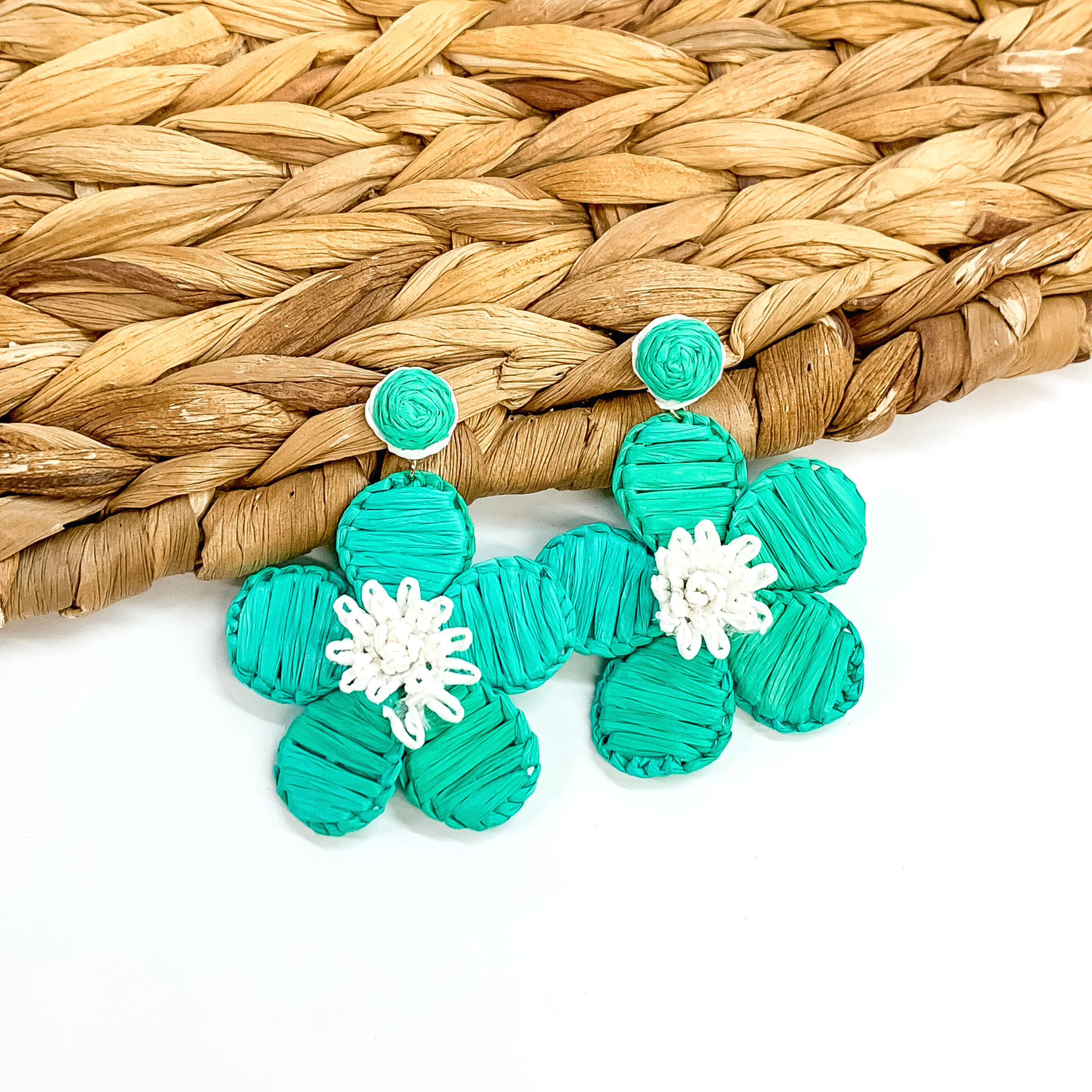 Turquoise raffia wrapped circle studs with hanging flower pendant wrapped in turquoise  raffia and white colored middle. These earrings are pictured partially laying on a basket weave material on a white background.