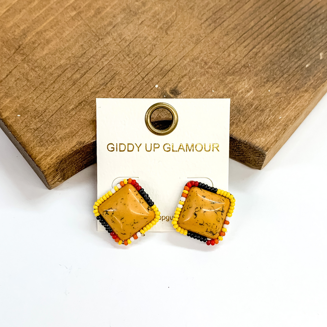 Yellow square stud earrings with a multicolored beaded outline. These earrings are pictured leaning against a brown block on a white background. 
