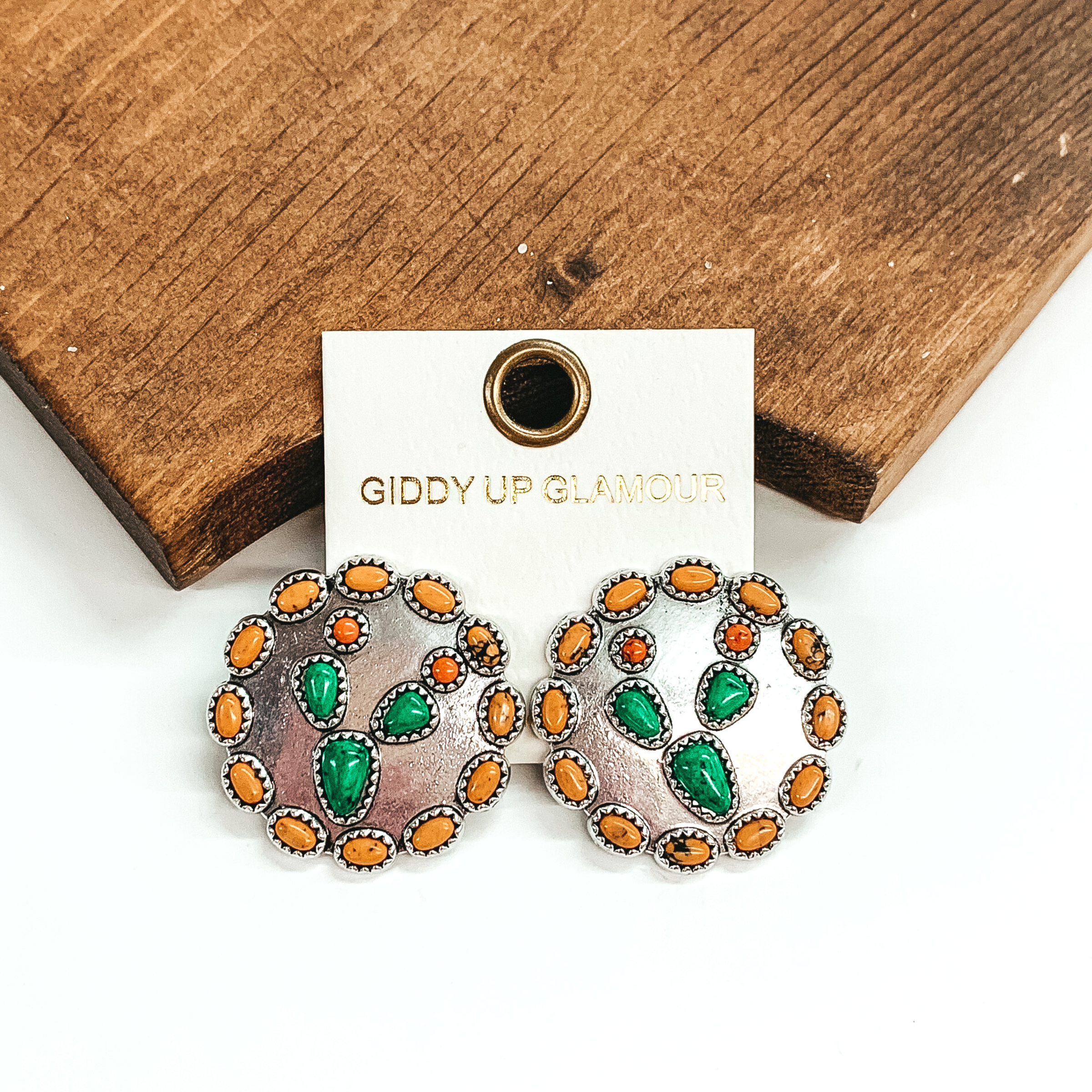 Silver circle earrings that are outlined in yellow stones and has a center green stone cactus. These earrings are pictured leaning against a brown block on a white background.