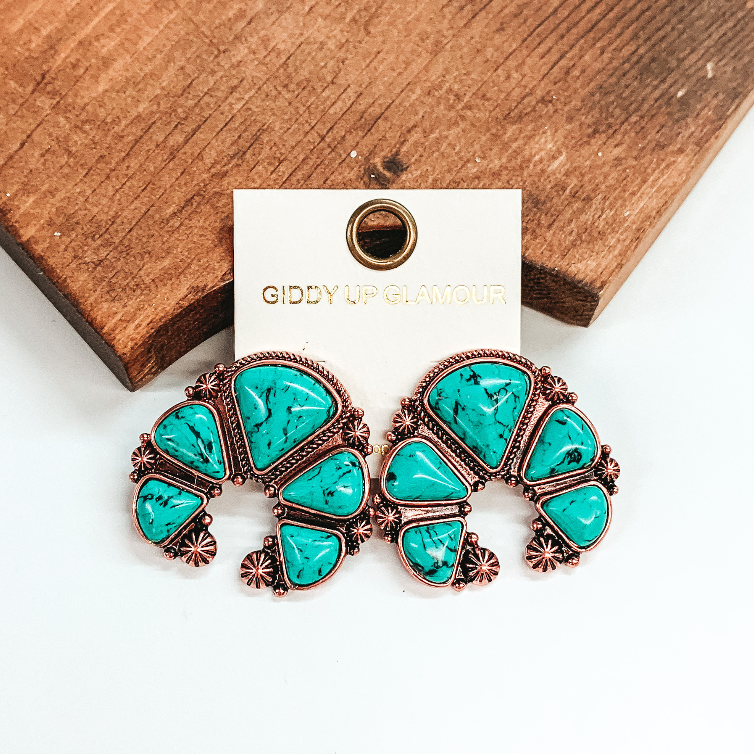 Copper naja earrings with turquoise stones. These earrings are pictured leaning against a brown block on a white background.
