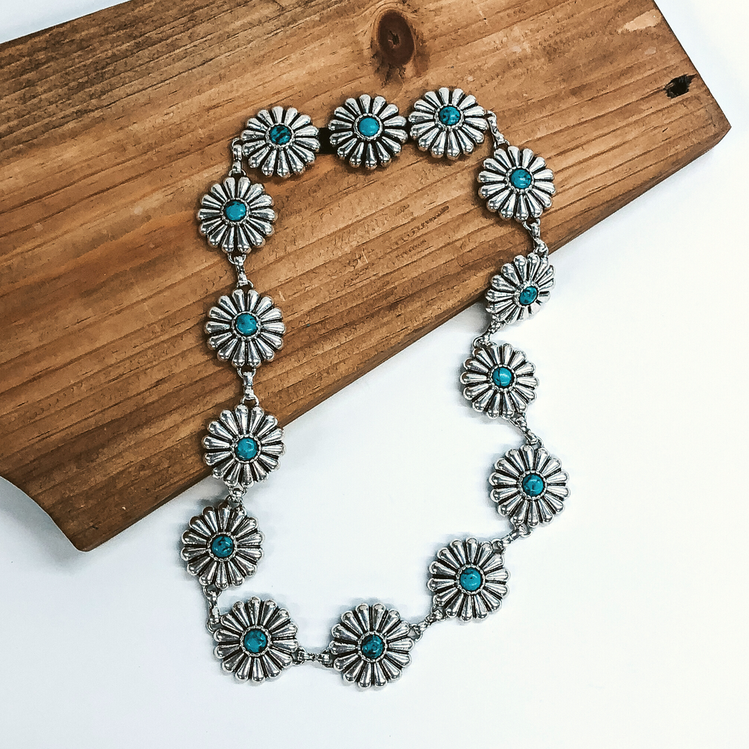 Silver, circle concho hat band with small turquoise stones in the middle of each concho. This hat band is pictured partially laying on a brown block on a white background.