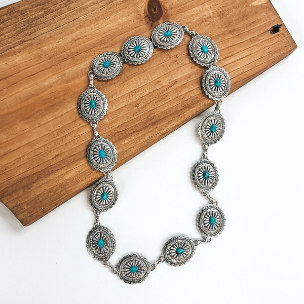 Silver, oval concho hat band with ab crystals and turquoise stones. This hat band is pictured partially laying on a brown block on a white background.