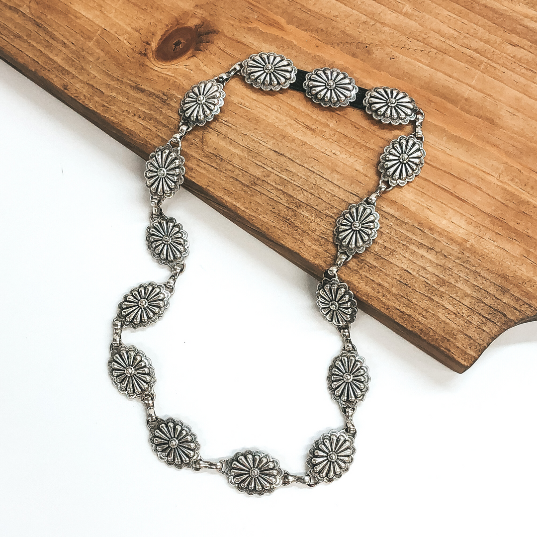 Silver, oval concho hat band. This hat band is pictured partially laying on a brown block on a white background.