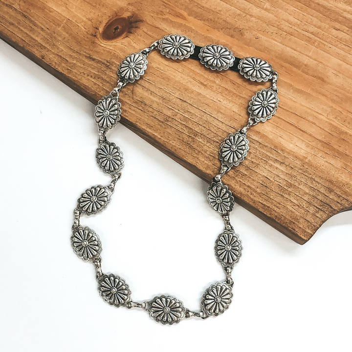 Silver, oval concho hat band. This hat band is pictured partially laying on a brown block on a white background.
