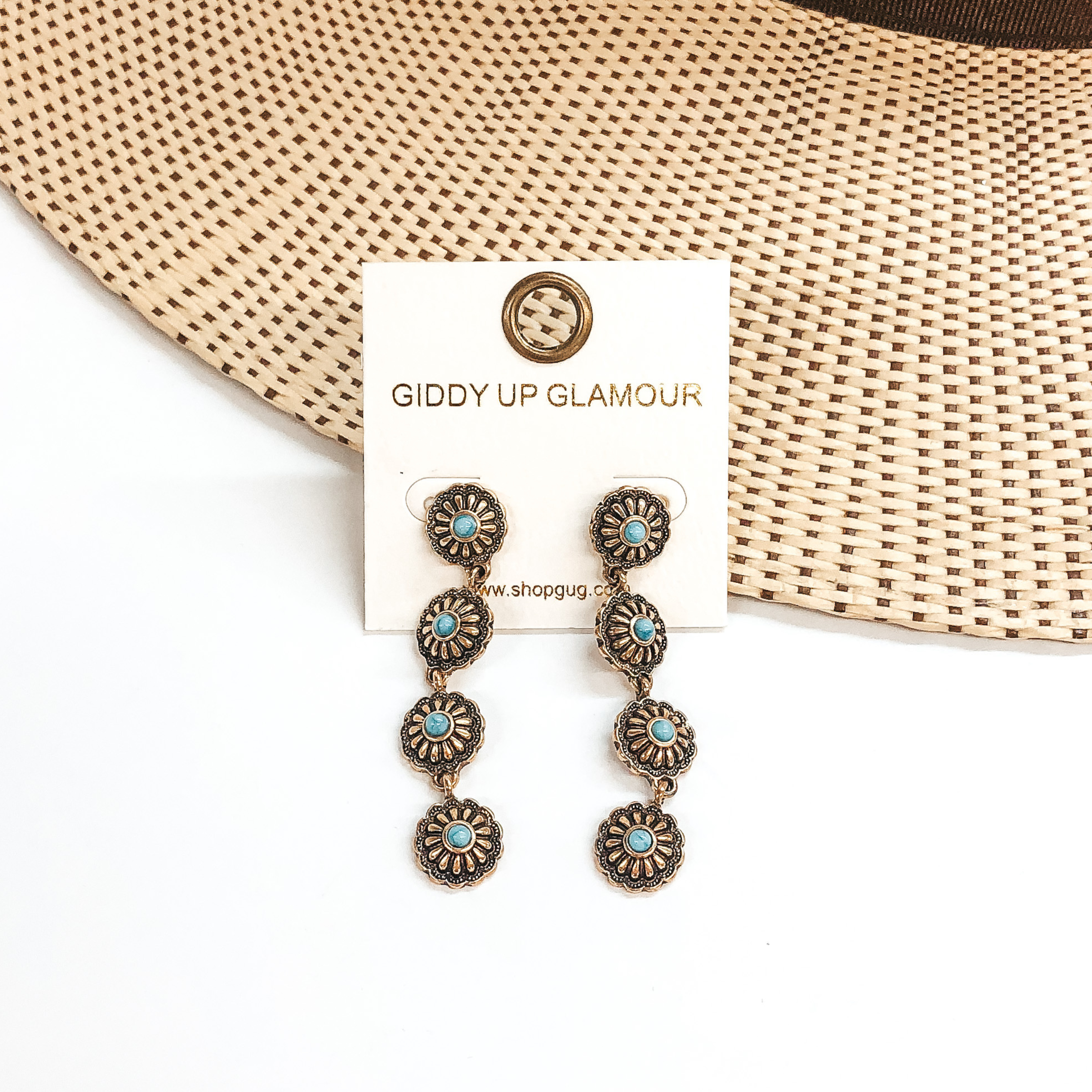 Four small, gold conchos in a row to form a dangle earrings. Each concho has a small turquoise stone in the center. These earrings are pictured on a white and tan background.