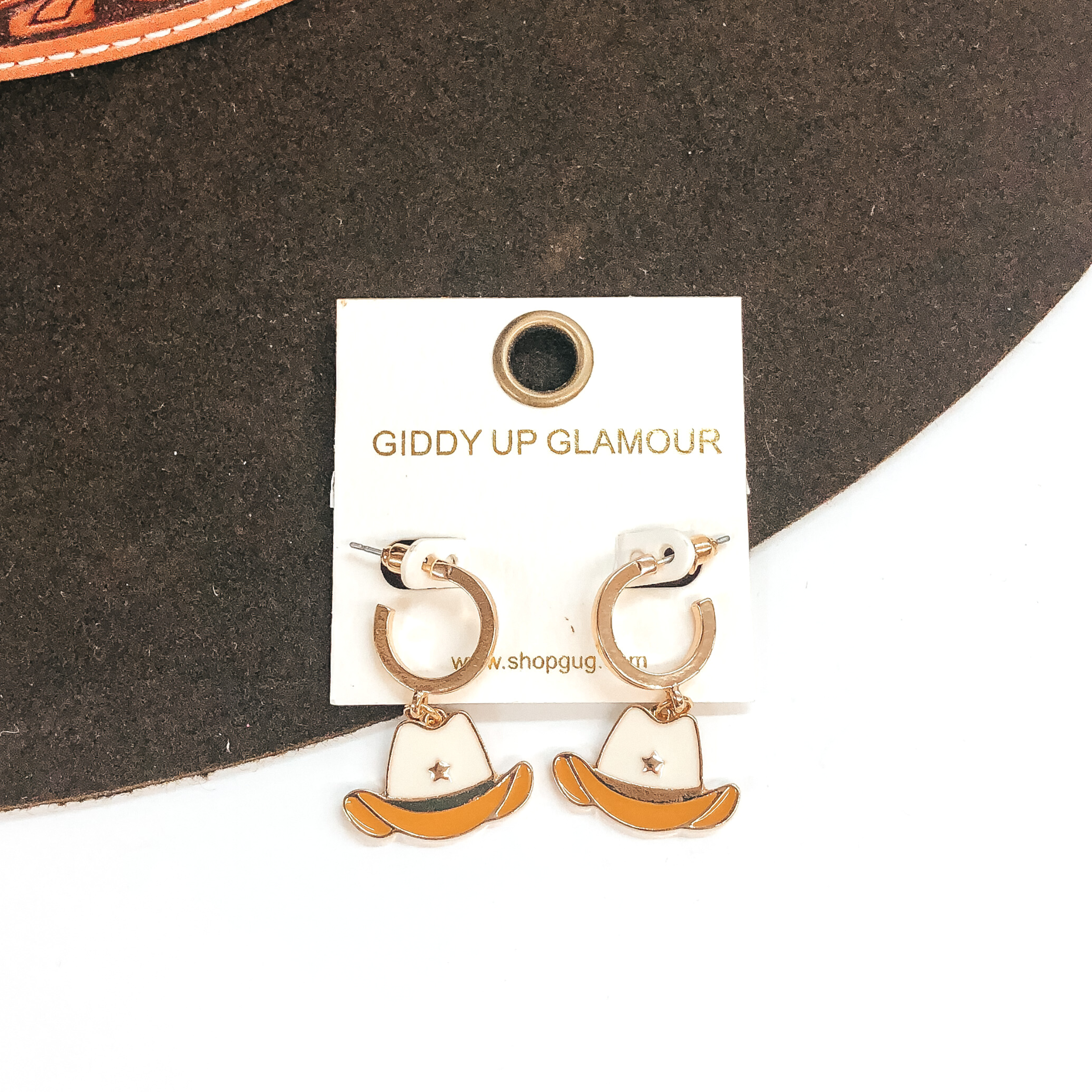Small gold hoop earrings with a ivory and tan hat pendant hanging from the bottom. These earrings are pictured on a white and brown background. 