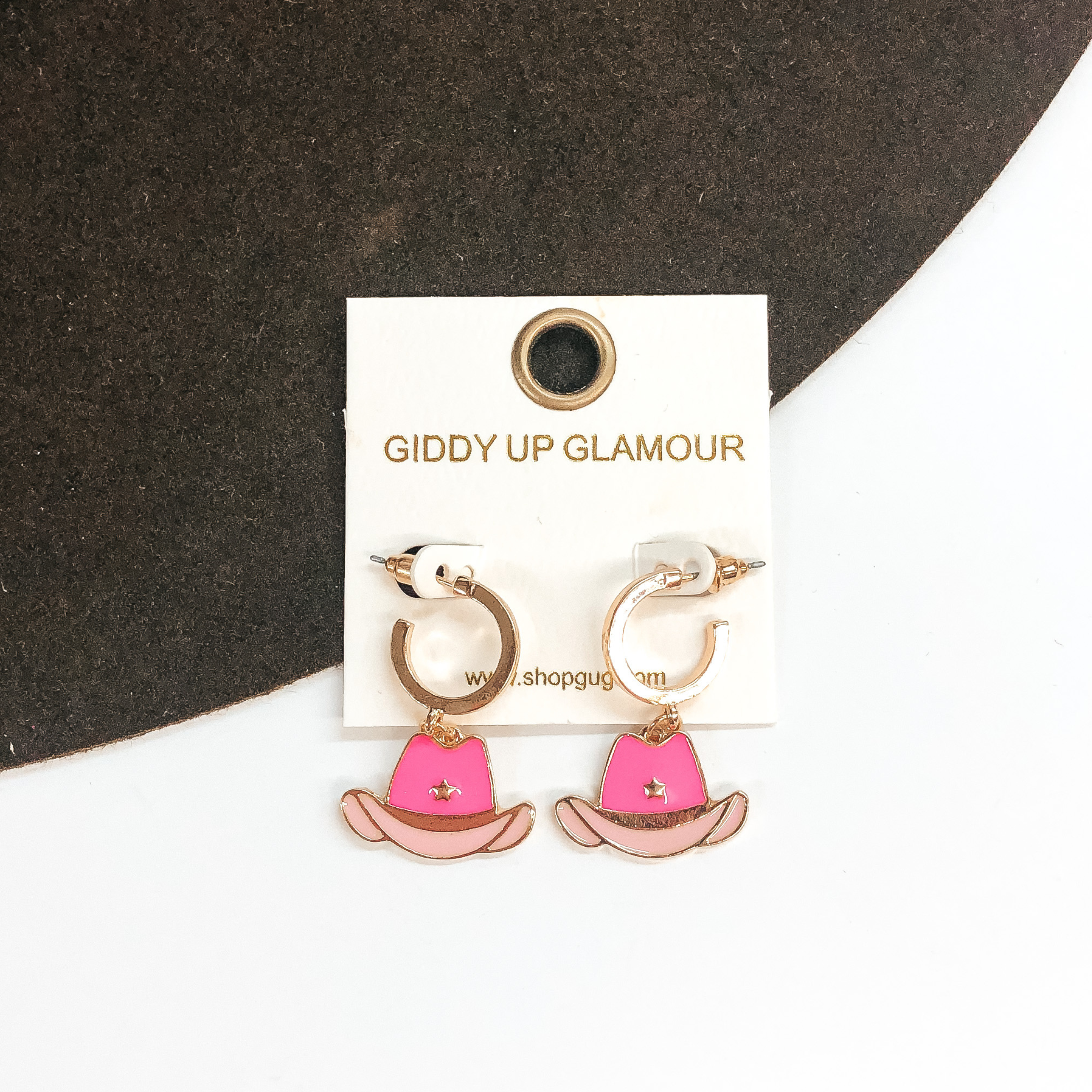 Small gold hoop earrings with a dark pink and light pink hat pendant hanging from the bottom. These earrings are pictured on a white and brown background. 