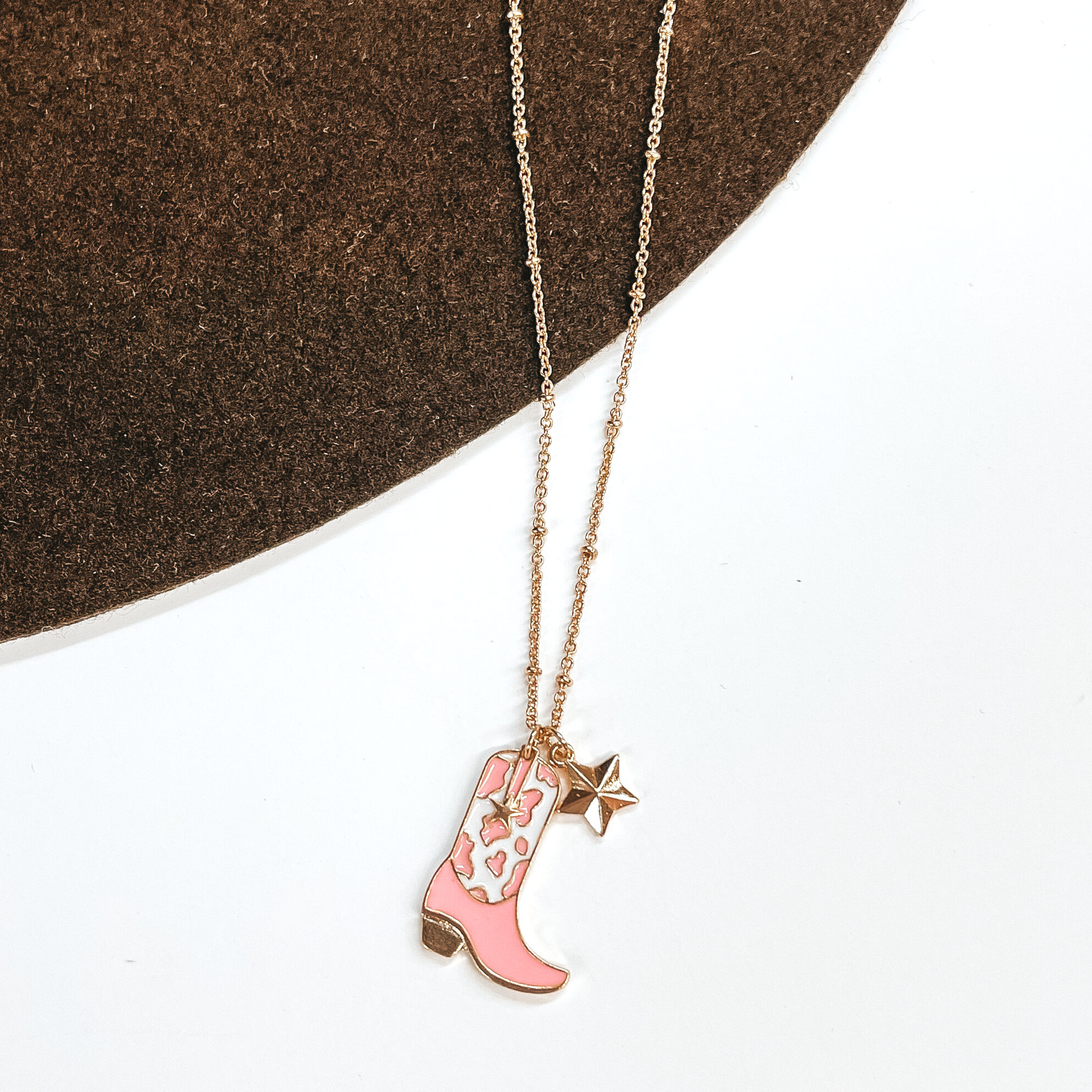 Gold chain necklace with a gold star charm and a white and pink cow print boot charm. This necklace is pictured on a white and brown background. 