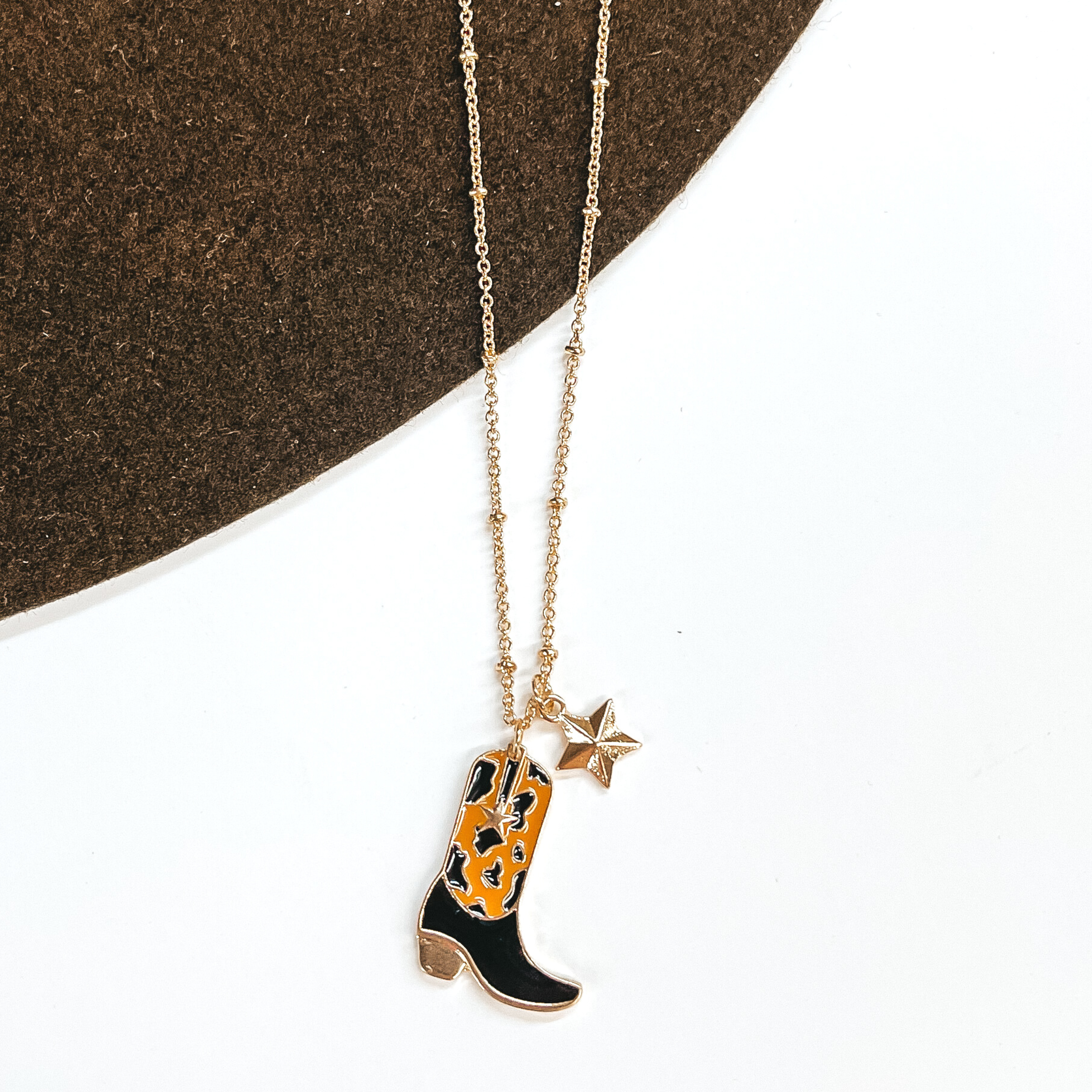Gold chain necklace with a gold star charm and a black and tan cow print boot charm. This necklace is pictured on a white and brown background.