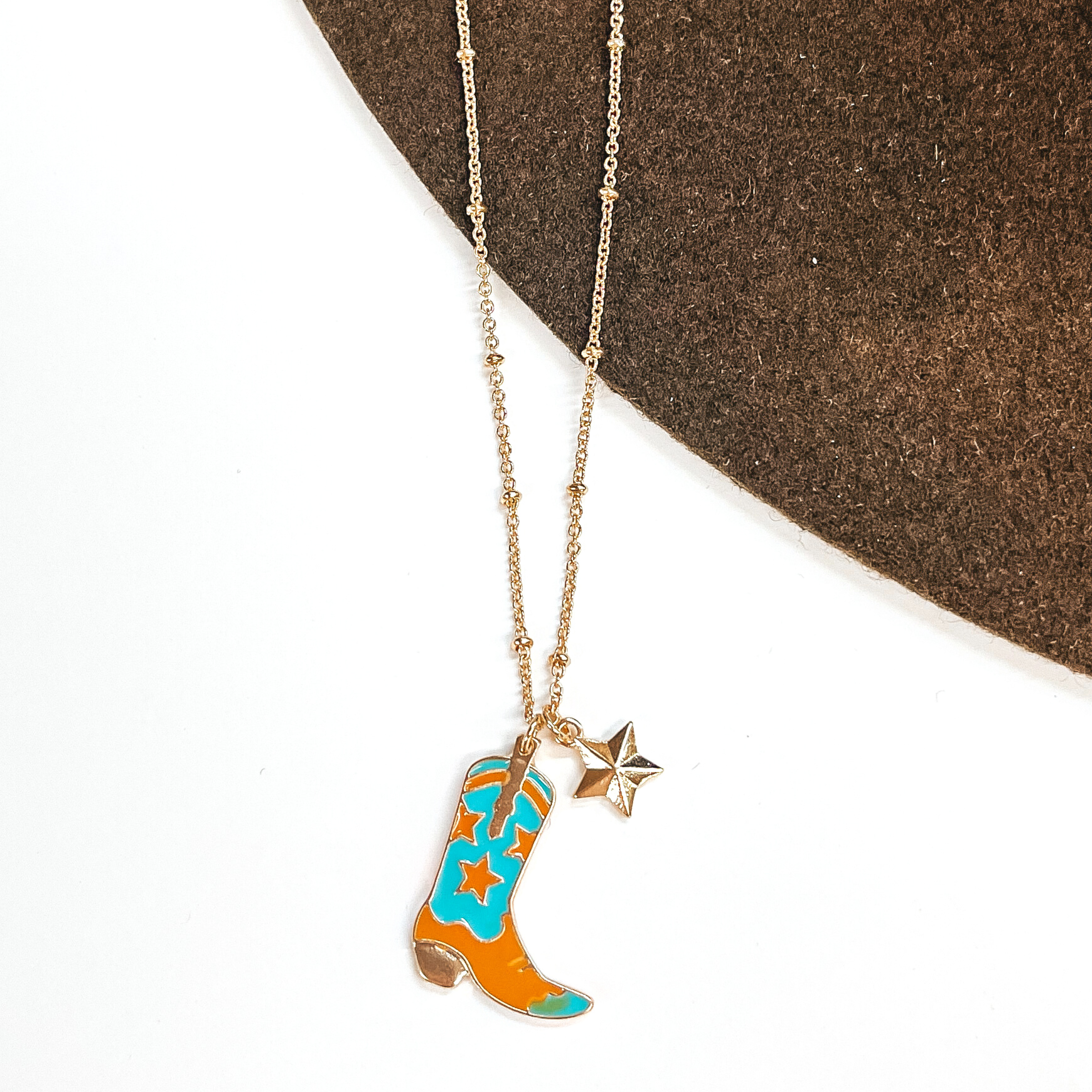 Gold chain necklace with a gold star charm and a turquoise and tan star print boot charm. This necklace is pictured on a white and brown background.