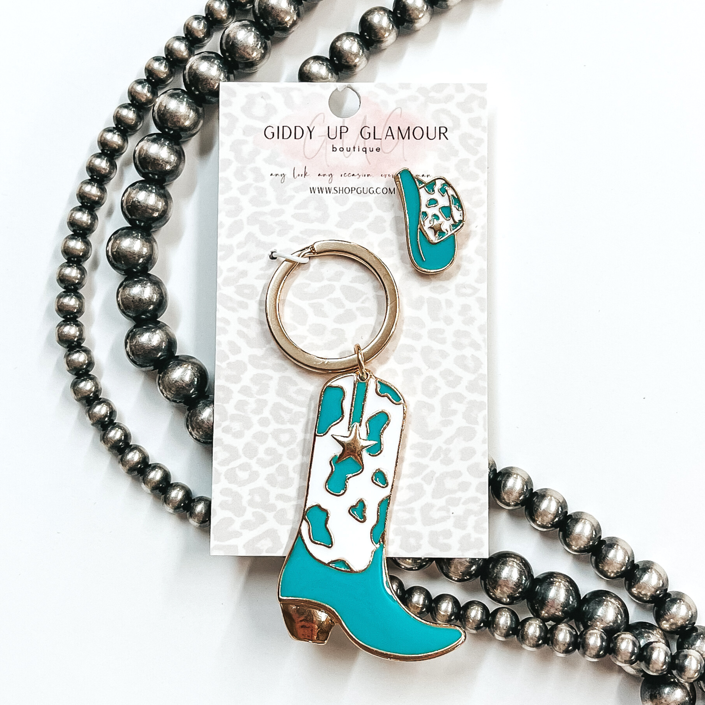 Gold key ring with boot pendant that has a white and turquoise cow print design. The boot also has a gold star. There is also a white and pink hat pin. These two are pictured on a white cardstock on a white background with silver beads. 