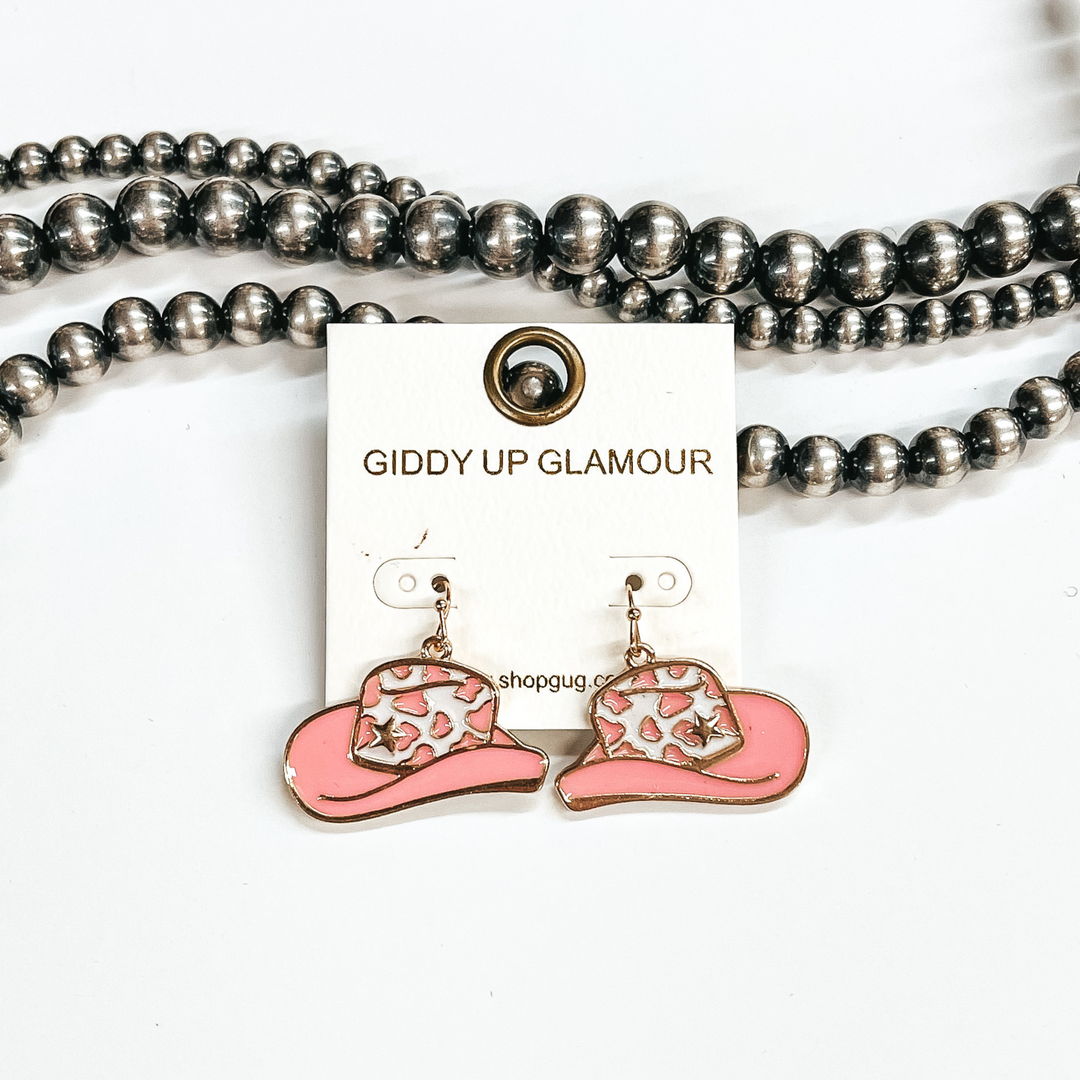 Gold fish hook earrings with hanging hat pendant. This hat is white and pink with a cow print design and gold star. These earrings are pictured on a white background with silver beads. 