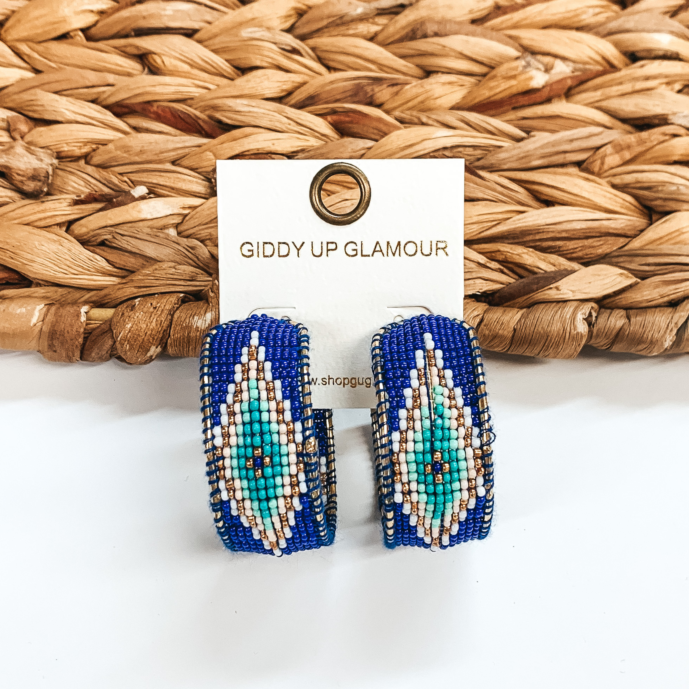 These beaded hoop earrings are mainly royal blue color with an Aztec pattern of white, light blue, and gold. These earrings are pictured on a white card holder in front of a basket weave material on a white background. 