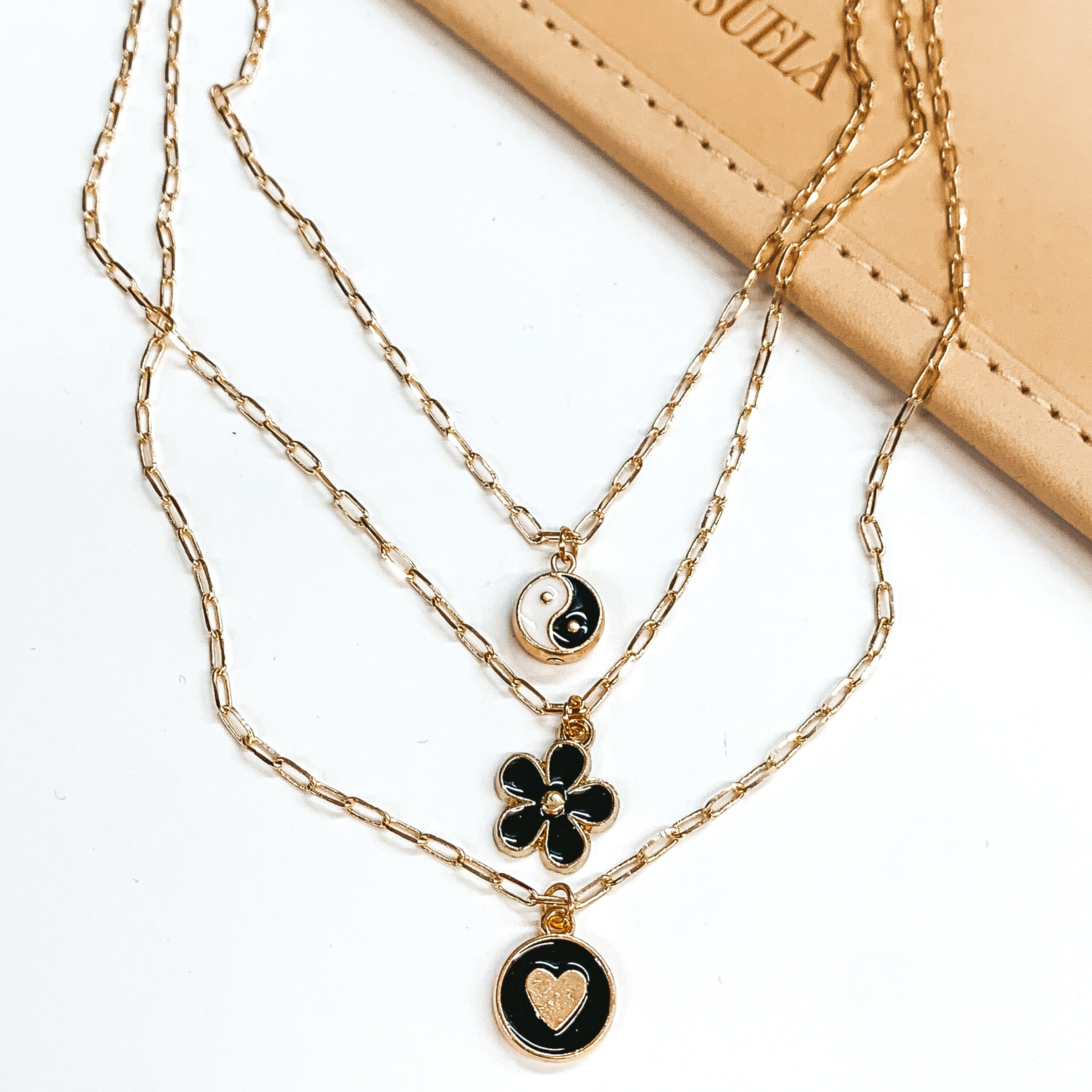 Three stranded gold chain necklace. Each strand has an individual charm in black, white, and/or gold. These include, a ying and yang circle charm, a flower charm, and a circle charm with a gold heart. This necklace is pictured partially on a tan bag on a white background. 