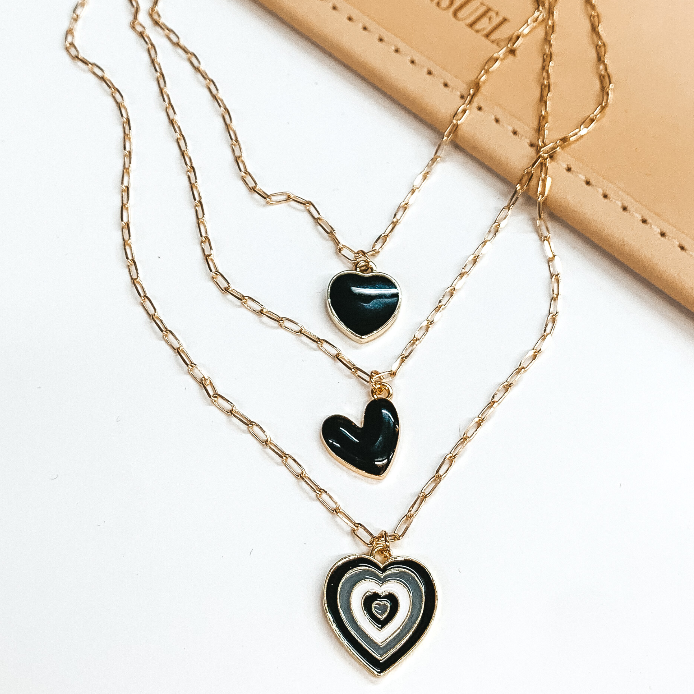 Three stranded gold chain necklace. Each strand has an individual charm in black, white, and/or gold. These include, three heart charms in different sizes. This necklace is pictured partially on a tan bag on a white background. 