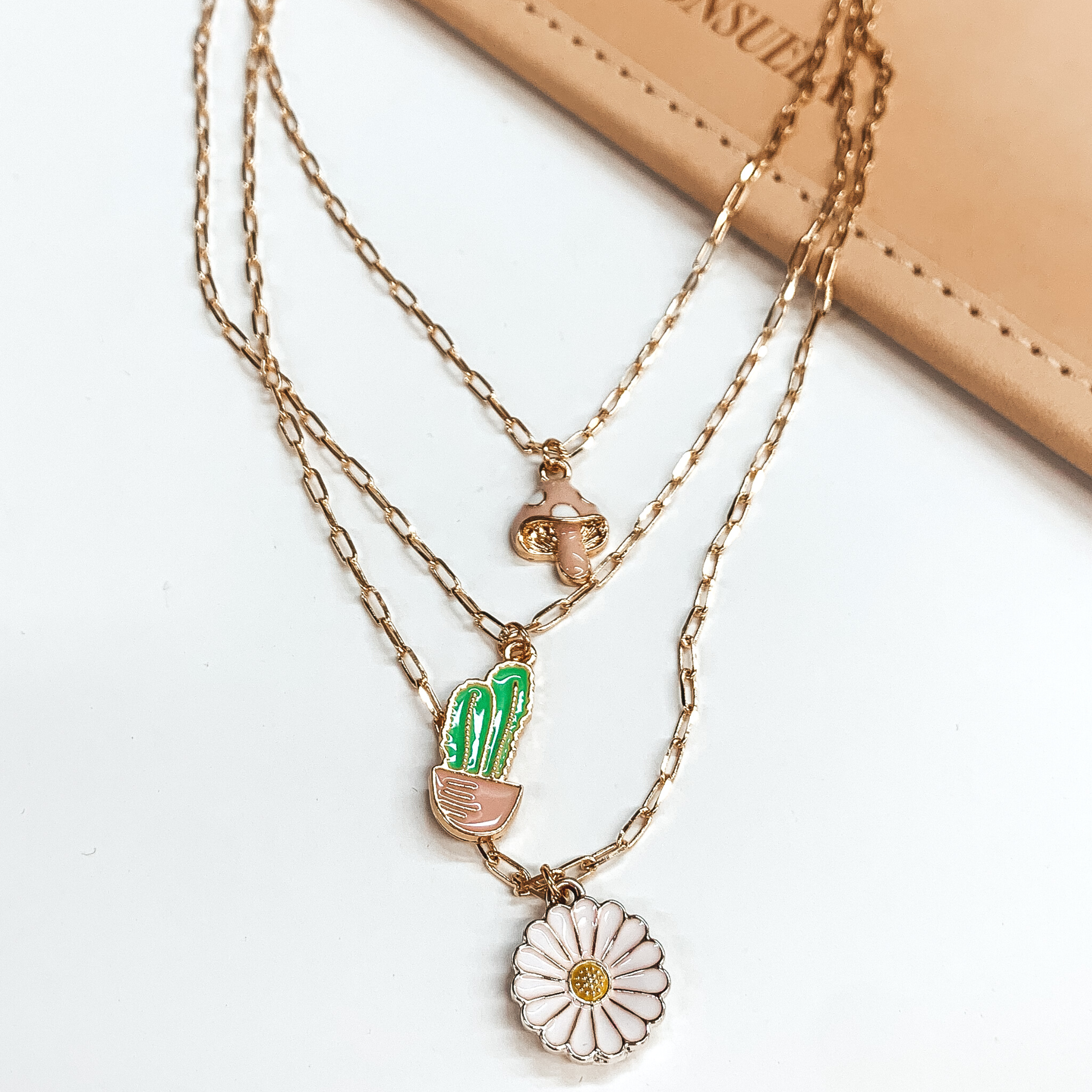 Three stranded gold chain necklace. Each strand has an individual charm in colors white, green, white, and pale pink. These include, a mushroom charm, a cactus in a pot charm, and a white flower charm. This necklace is pictured partially on a tan bag on a white background. 