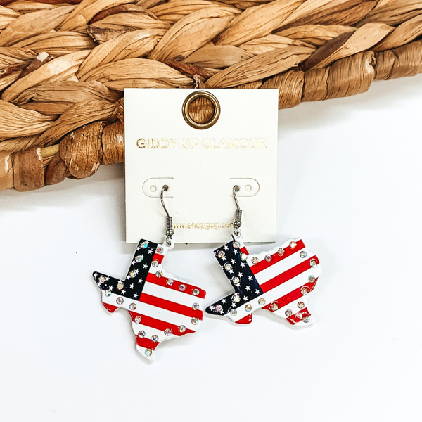 Silver fish hook earrings with a texas shaped pendant. This pendant has an american flag design with an AB crystal outline. These earrings are pictured in front of a basket weave material on a white background. 