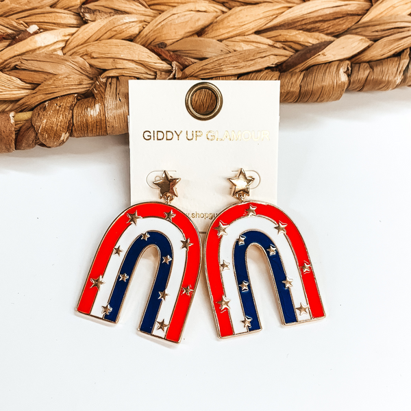 Gold star post earrings with a hanging arch. The arch has a red, blue, and white stripe with gold stars throughout. These earrings are pictured in front of a basket weave material on a white background. 
