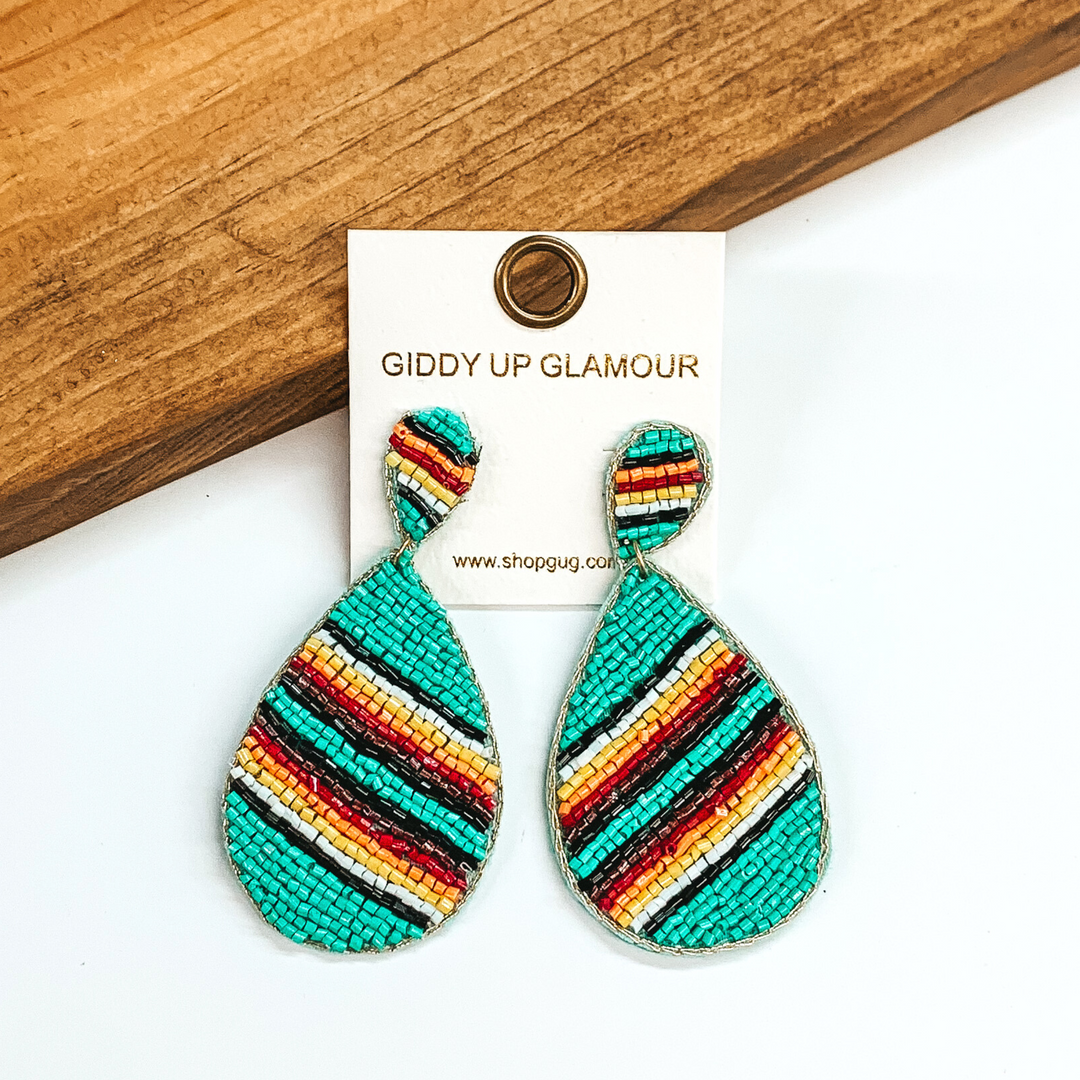 Beaded upside down teardrop stud earrings with a hanging teardrop. The main color of the beads is turquoise with multicolored stripes. These earrings are pictured on a white and brown wood background. 