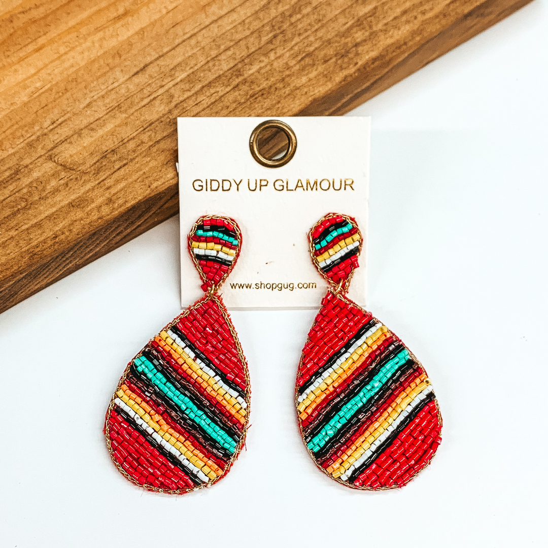 Beaded upside down teardrop stud earrings with a hanging teardrop. The main color of the beads is red with multicolored stripes. These earrings are pictured on a white and brown wood background.