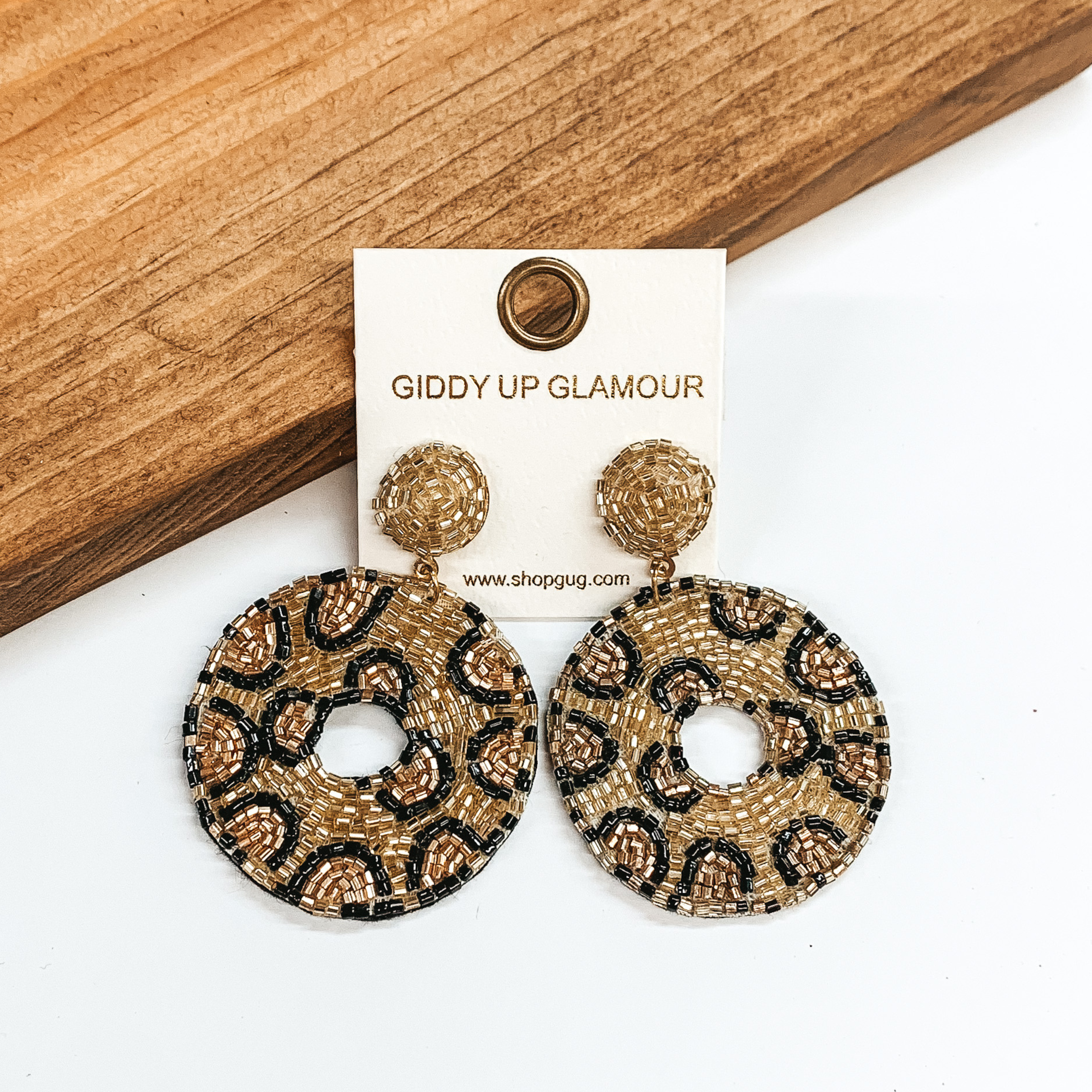 White circle beaded post back earrings in gold. These earrings include a beaded drop circle in gold with a leopard print. These earrings are pictured on a white and brown wood background.