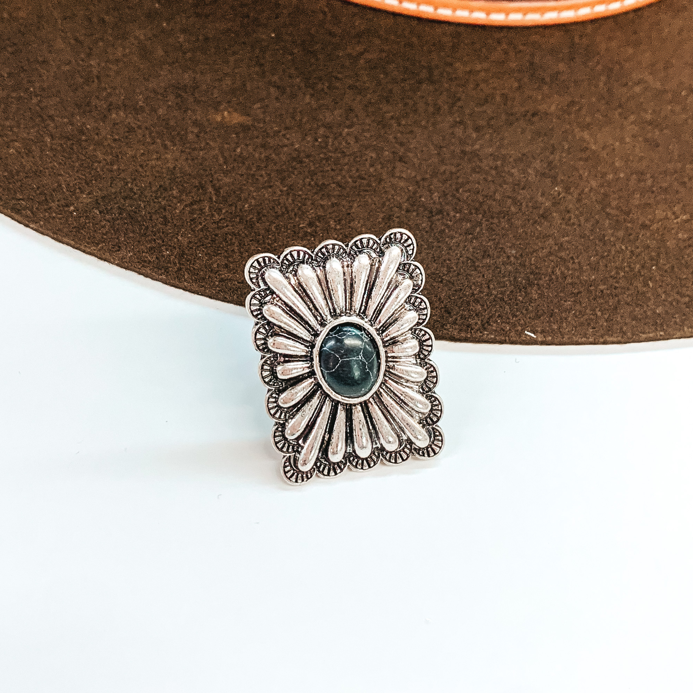 Silver, square concho ring with a center black stone. This ring is pictured in front of a brown hat on a white background.