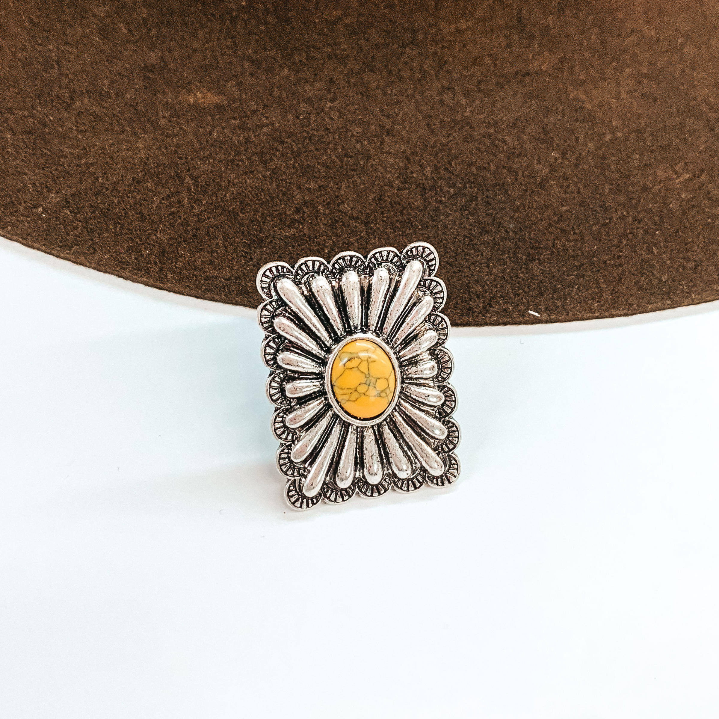 Silver, square concho ring with a center yellow stone. This ring is pictured in front of a brown hat on a white background.