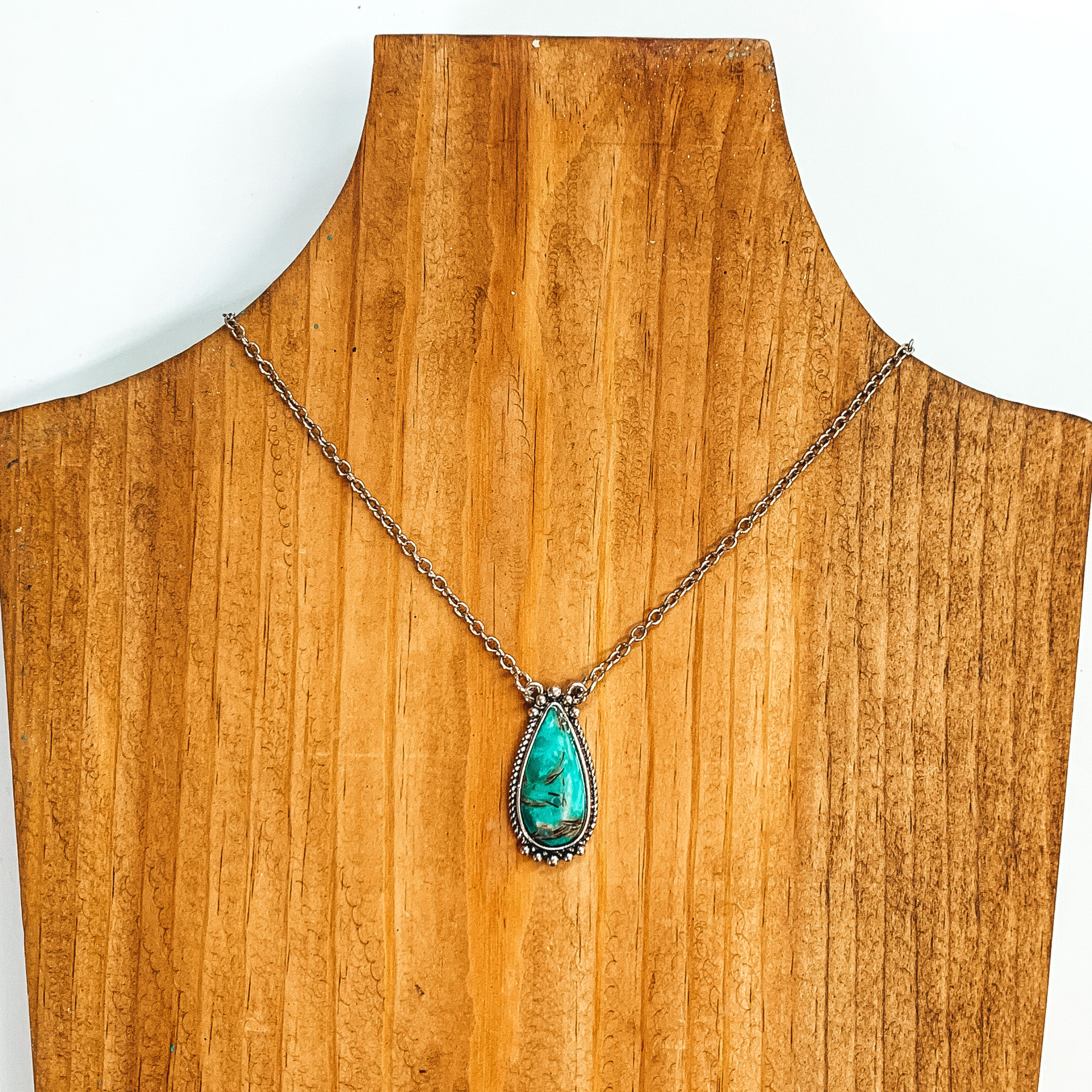 Silver chain necklace with a turquoise teardrop pendant with a silver outline. This necklace is pictured on a wood necklace holder on a white background. 