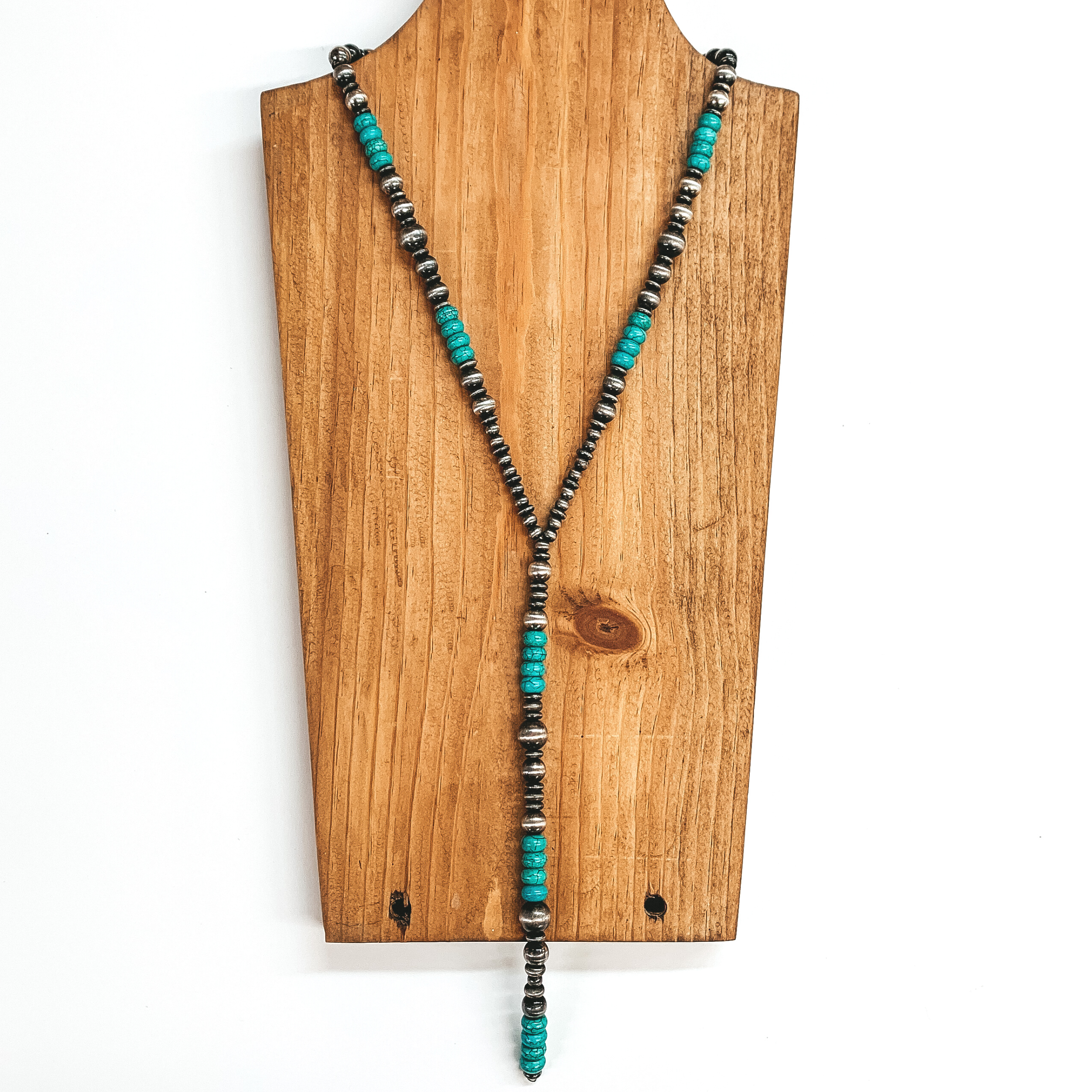 Silver and turquoise beaded necklace with a long beaded drop. The silver beads have different sizes of beads, while the turquoise beads are the space size and used as spacers. This necklace is pictured on a wood necklace holder on a white background. 
