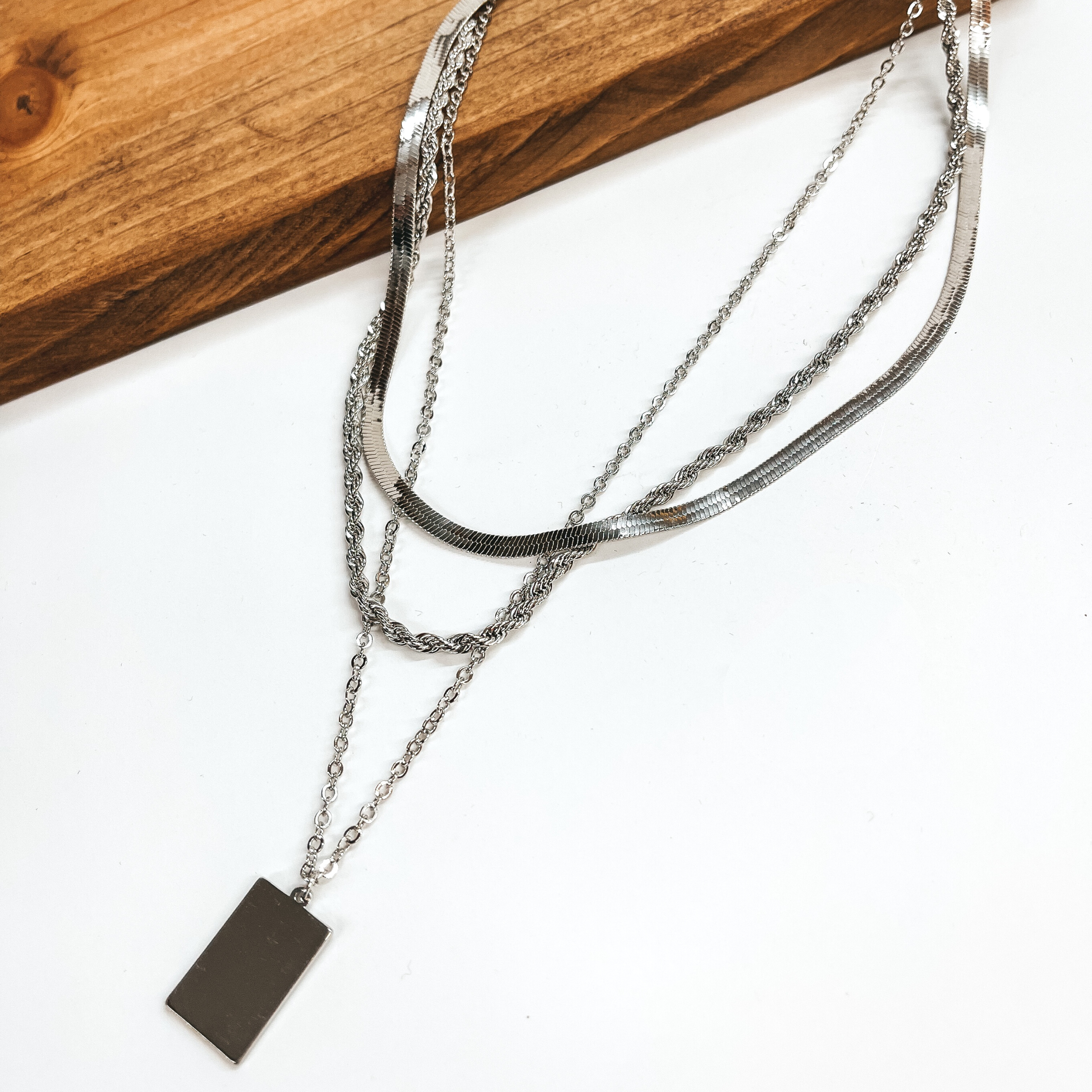Silver chain necklaces pictured partially laying on a wood block with the rest of the necklaces on a white background. This pictures includes a snake chain, a longer rope chain, and an even long regular chain with a silver rectangle pendant. 
