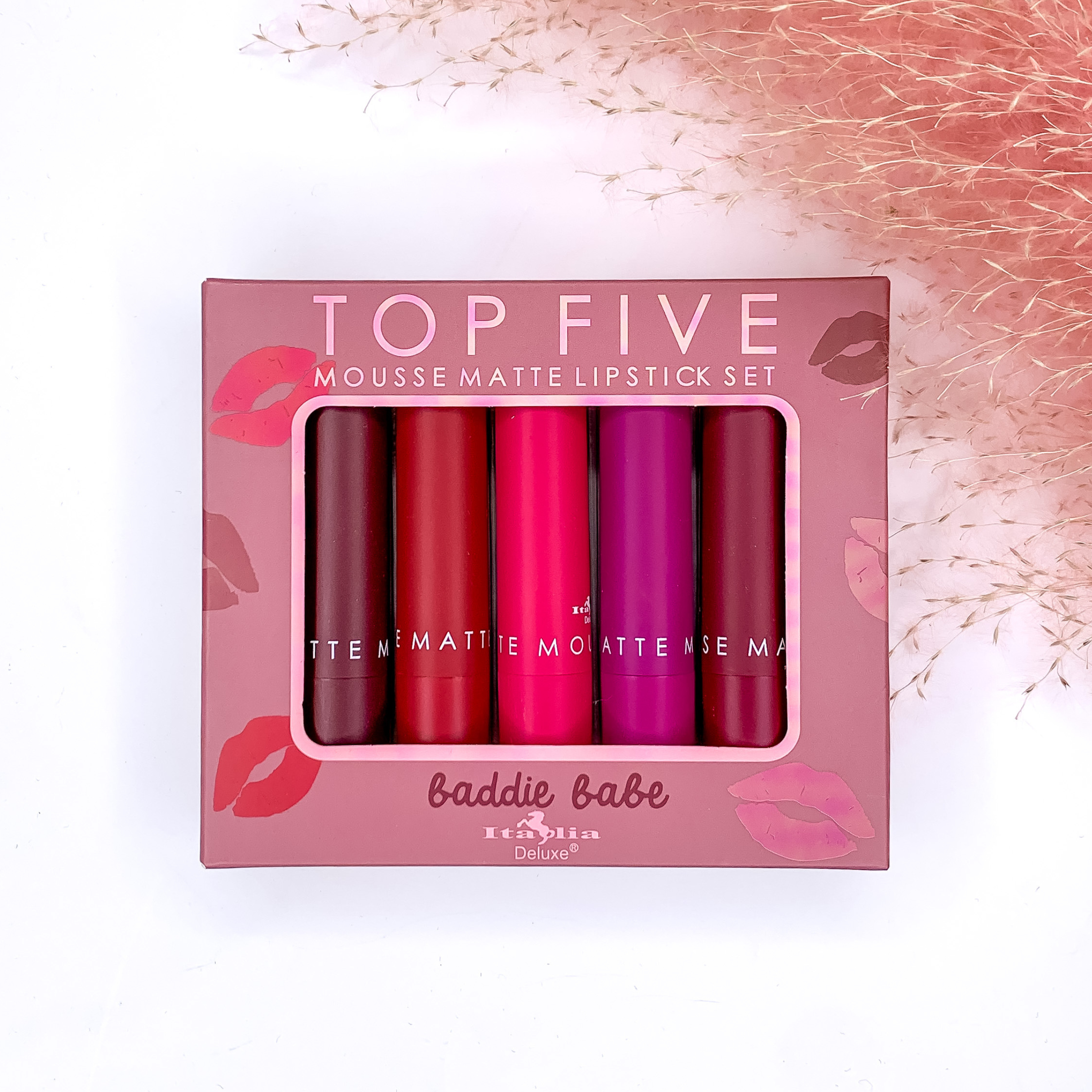 In a light purple box, there are 5 lip stick tubes. Colors included are dark purple, red, pink, purple, and dark red. This set is pictured on a white background with pink pompus grass in the top right corner. 