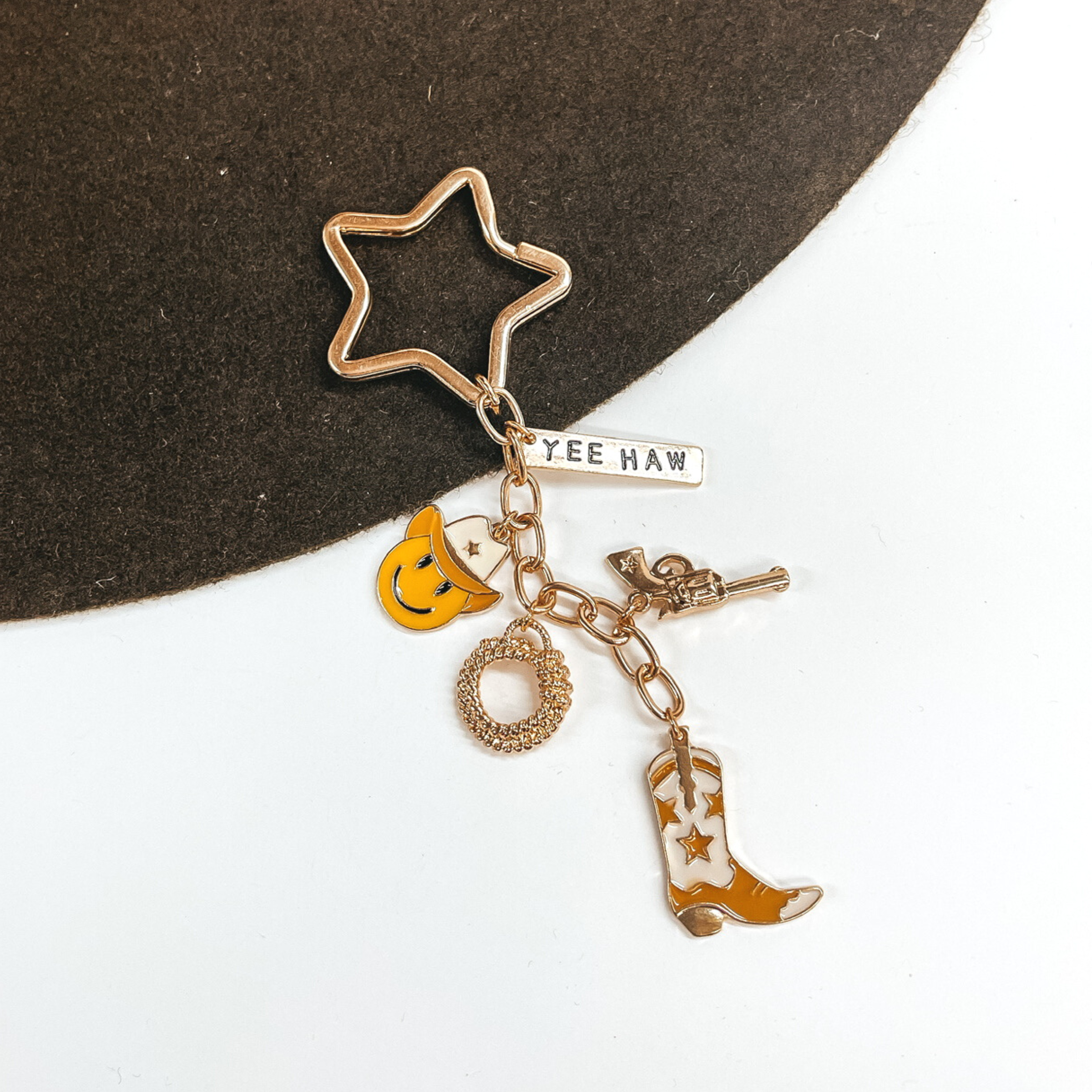 Gold, star shaped key ring with a hanging gold chain with charms on it. There is a gold rectangle charm with the words "YEE HAW," a gold gun charm, a gold rope charm, a yellow smiley face chram with a ivory cowboy hat, and an ivory and tan cowboy boot charm. This is pictured on a brown and white background. 