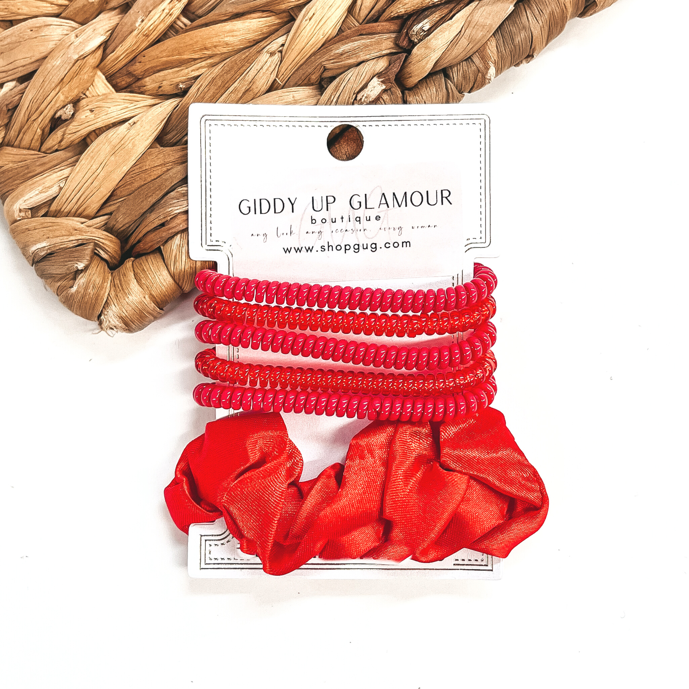 A set of six hair ties, five spiral and one satin scrunchie in red. There are three  solid color spiral hair ties and two transparent. These hair ties are on a white  cardboard piece with a Giddy Up Glamour logo, they are leaned up against a brown woven  plate and a white background.