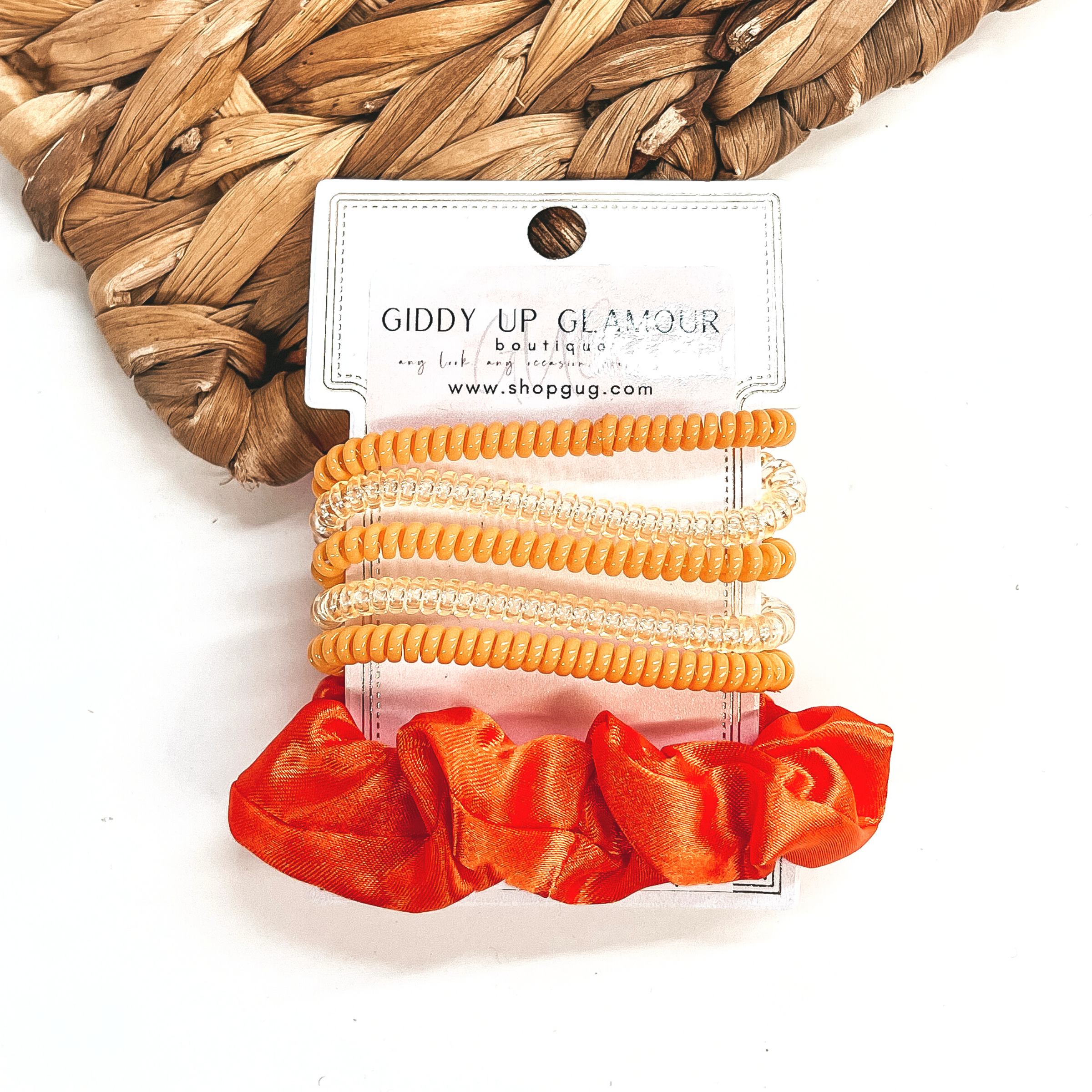 A set of six hair ties, five spiral and one satin scrunchie in orange. There are three  solid color spiral hair ties and two transparent. These hair ties are on a white  cardboard piece with a Giddy Up Glamour logo, they are leaned up against a brown woven  plate and a white background.