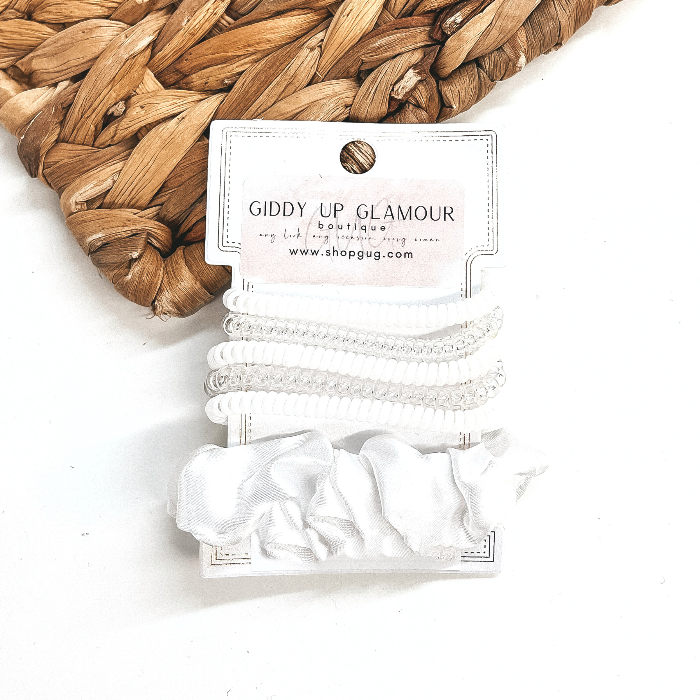 A set of six hair ties, five spiral and one satin scrunchie in white. There are three  solid color spiral hair ties and two transparent. These hair ties are on a white  cardboard piece with a Giddy Up Glamour logo, they are leaned up against a brown woven  plate and a white background.